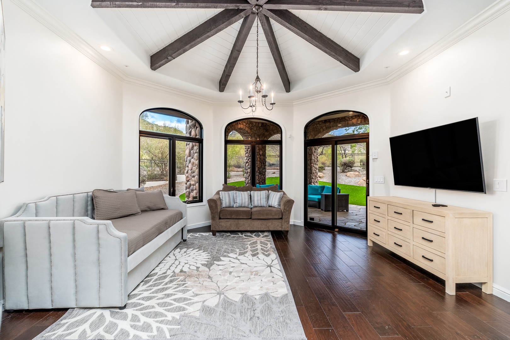 Sink into plush sofas beneath soaring vaulted ceilings with rustic wood beams. Sunlight floods through arched doors, framing your view of the lush backyard oasis beyond.