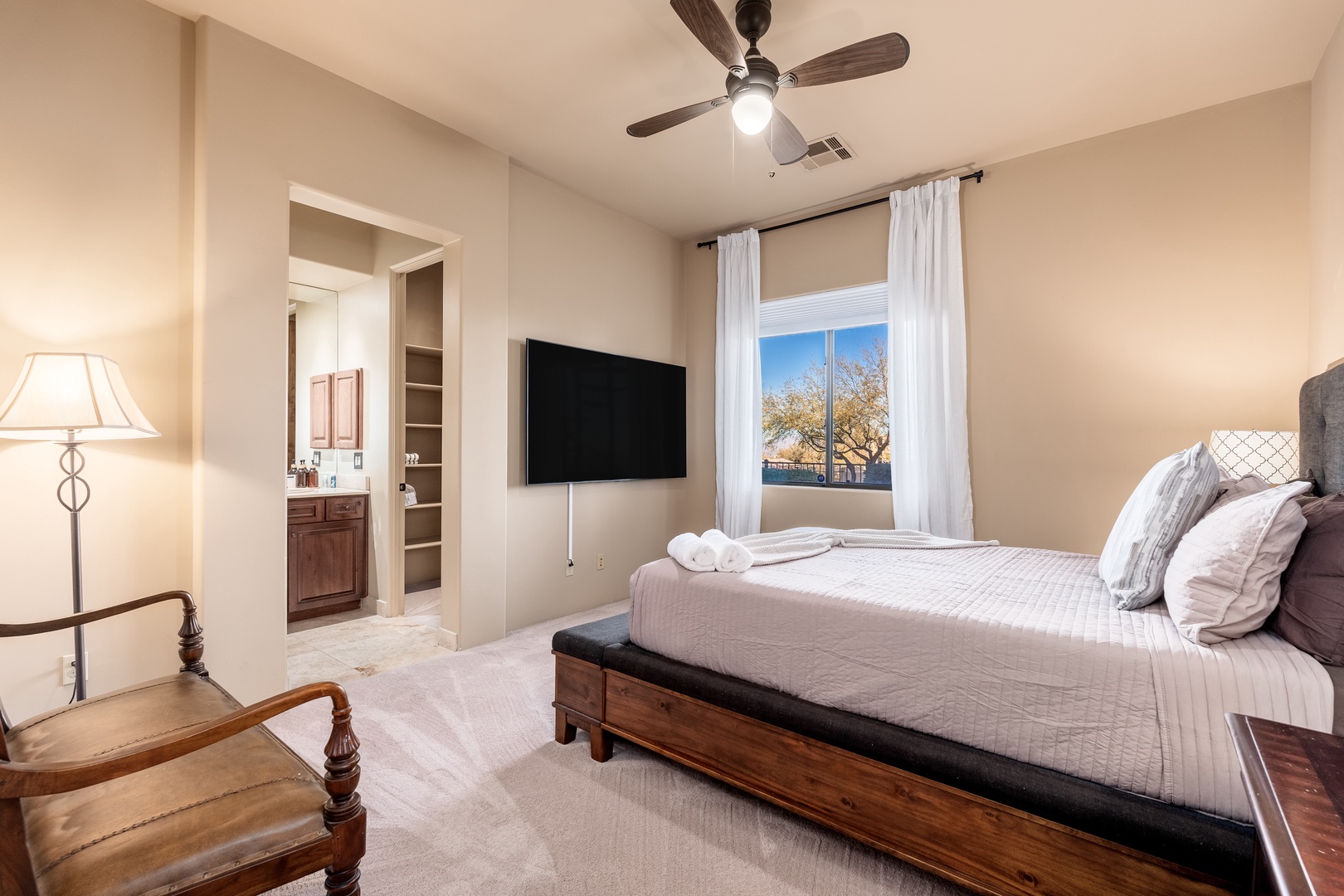 Unwind in your serene bedroom featuring a plush queen bed, flat-screen TV, and en-suite bathroom. The ceiling fan keeps you comfortable while natural light filters through the window, framing peaceful views.