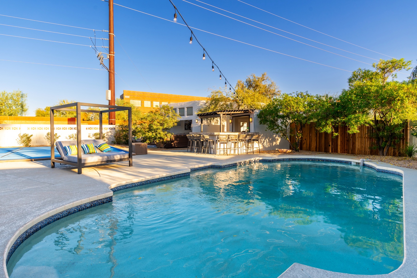 Modern vacation property featuring a sparkling pool, shaded cabana with lounge seating, outdoor bar area, and vibrant landscaping in a private desert setting.