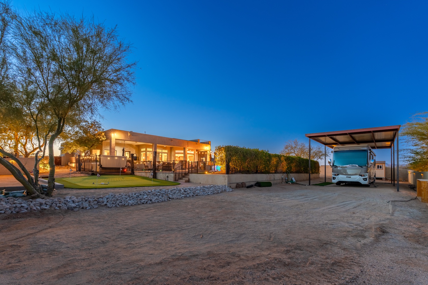 Modern desert property illuminated at twilight, with covered RV parking and landscaped grounds beneath a clear evening sky.