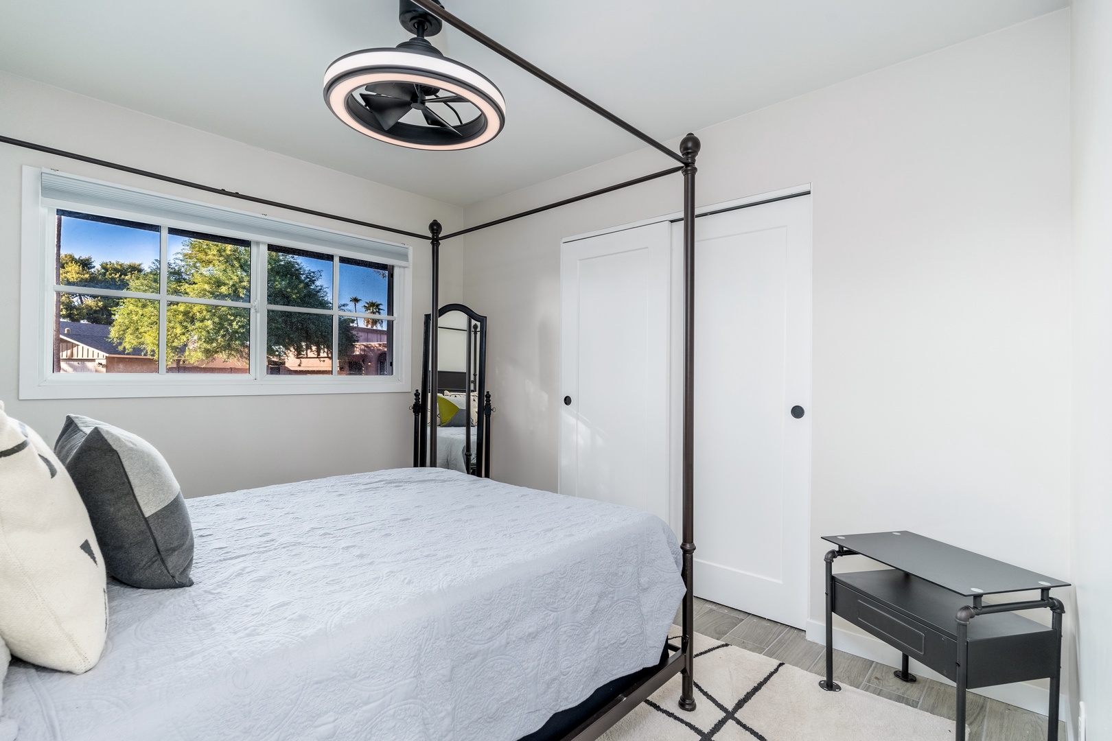 Your bright, peaceful bedroom features a striking canopy bed and modern ceiling fan, with large windows offering views of lush greenery outside—perfect for restful mornings.