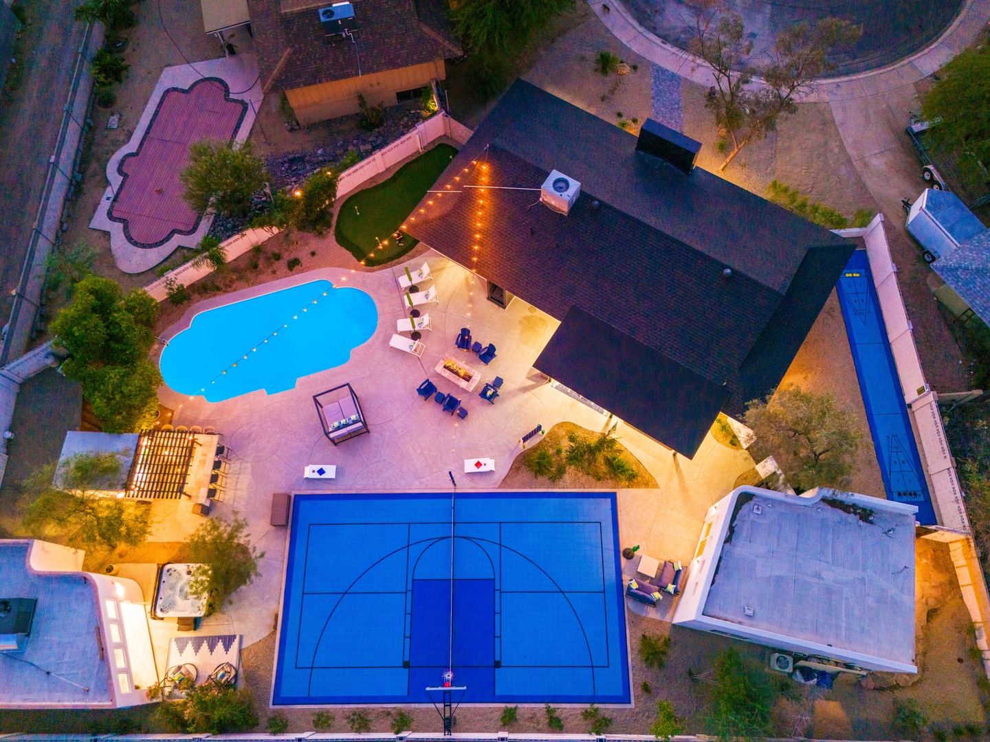 Aerial view of a private vacation rental featuring a full-sized basketball court, resort-style pool, outdoor dining pavilion, hot tub, and multiple entertainment areas surrounded by lush landscaping and desert scenery.