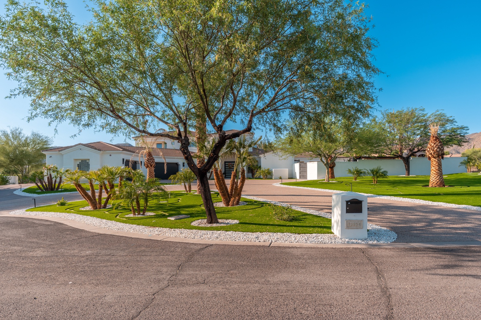 A spacious desert property featuring lush landscaping, mature trees, palm accents, and mountain views. The gated entrance and manicured grounds create a welcoming arrival experience.