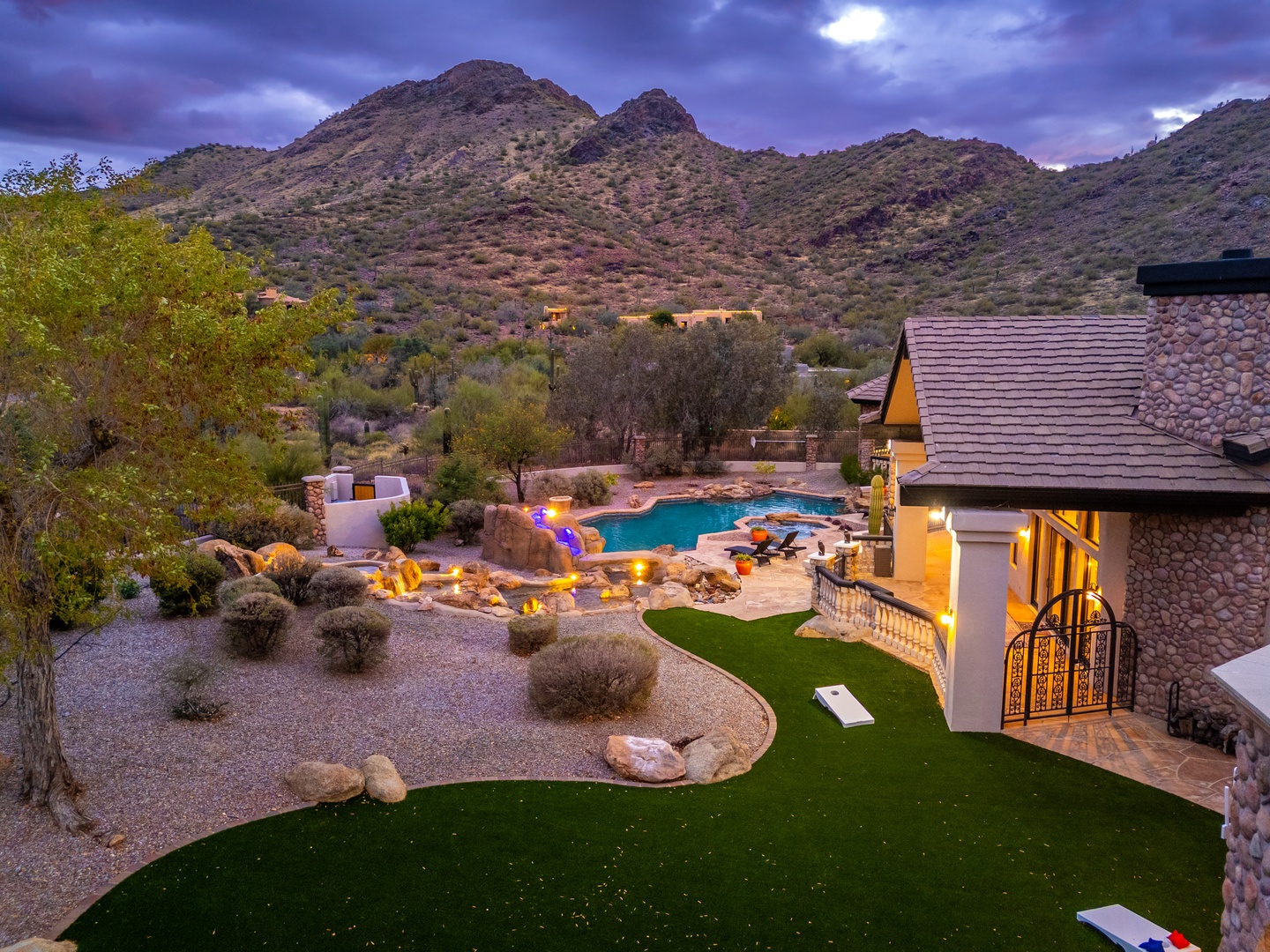 Twilight illuminates a stunning desert estate nestled against rugged mountain peaks, featuring a sparkling pool, lush landscaping, and elegant outdoor lighting throughout the grounds.