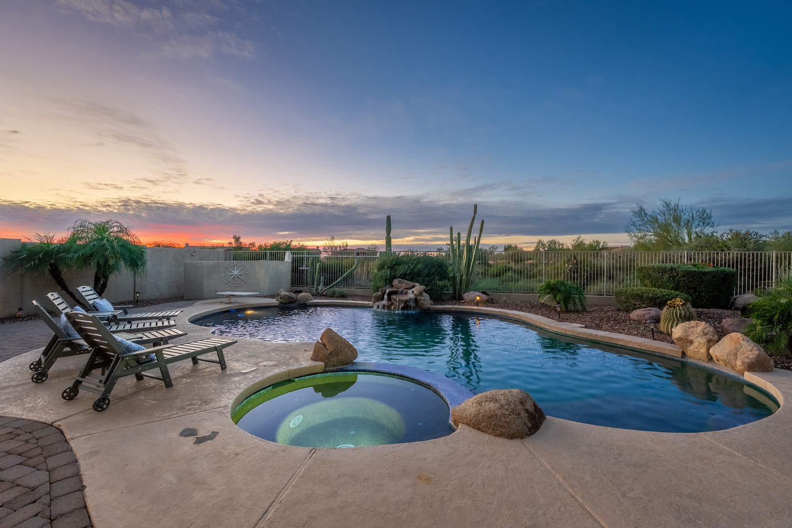 A desert-landscaped backyard featuring a freeform pool with rock waterfall, integrated spa, and loungers set against a stunning Arizona sunset and mountain views.