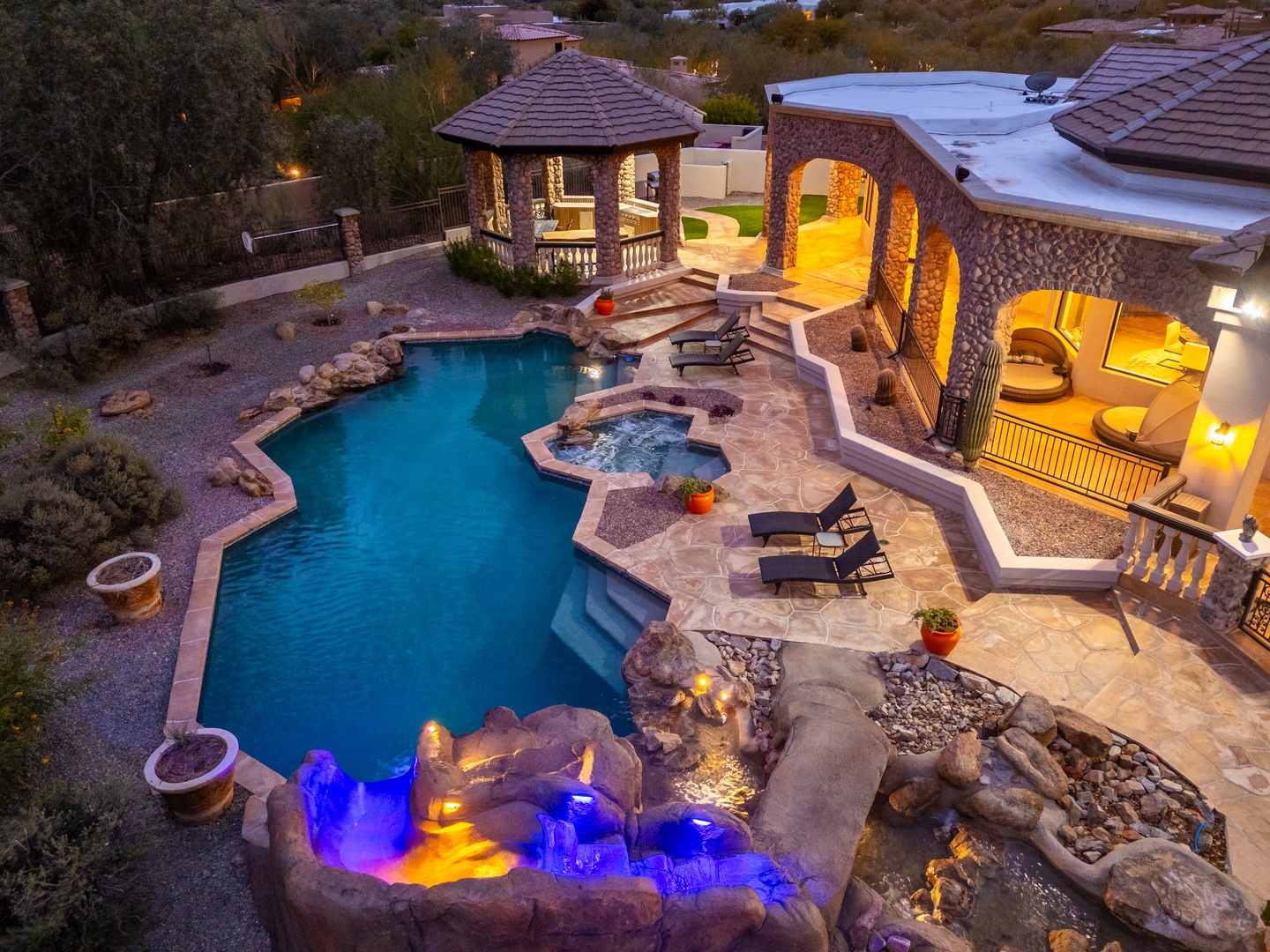 Aerial view of the property at dusk showcasing a stone-facade exterior, private pool with integrated hot tub and cascading waterfall feature, covered gazebo, illuminated arched outdoor living spaces, and surrounding desert landscape dotted with homes and 