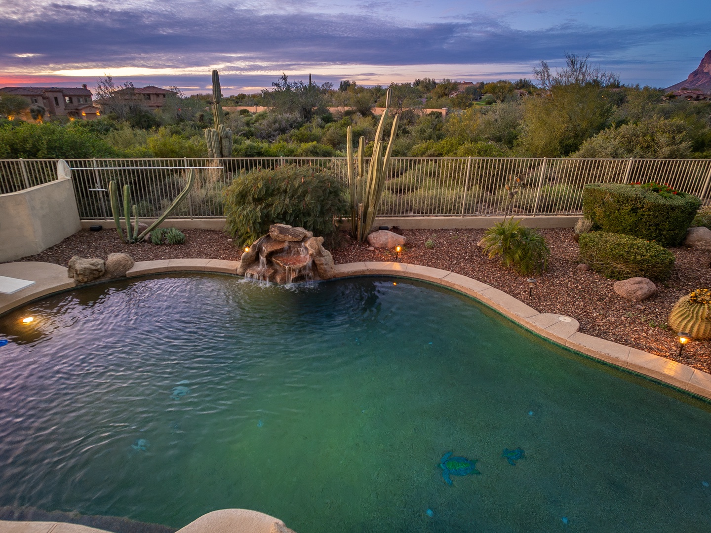 A stunning desert retreat featuring a private pool overlooking Sonoran landscape, with natural rock waterfall, saguaro cacti, and dramatic sunset skies painting the horizon—perfect for peaceful Arizona evenings.