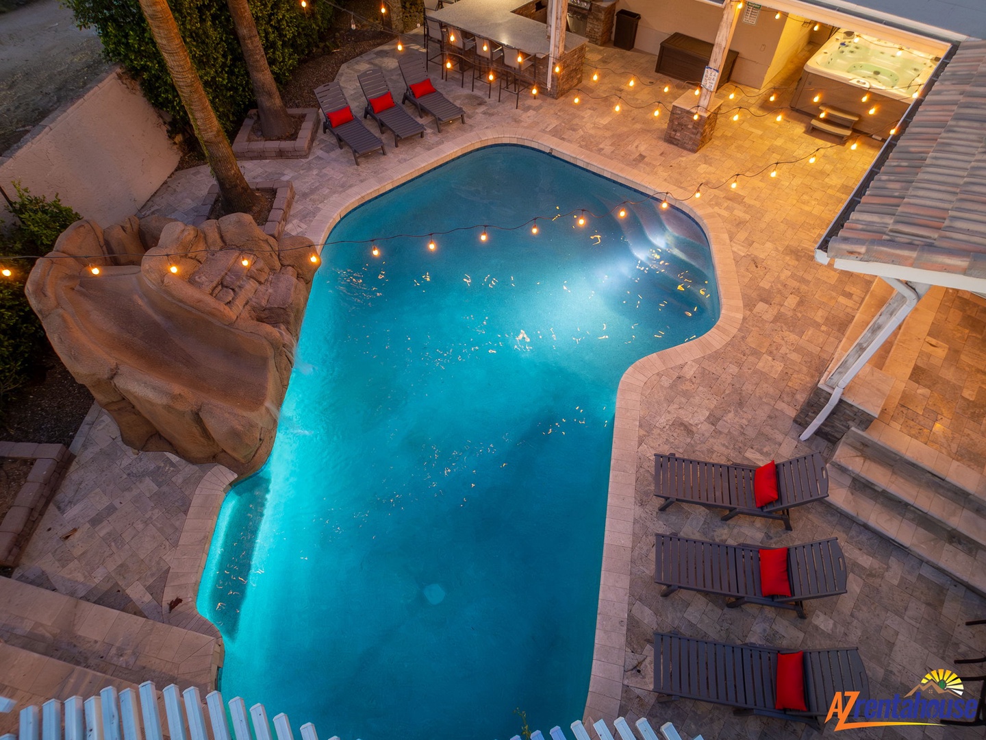 An aerial view captures the property's outdoor area, featuring a uniquely shaped pool surrounded by paver deck, string lights overhead, and loungers with vibrant red accents creating an inviting atmosphere.