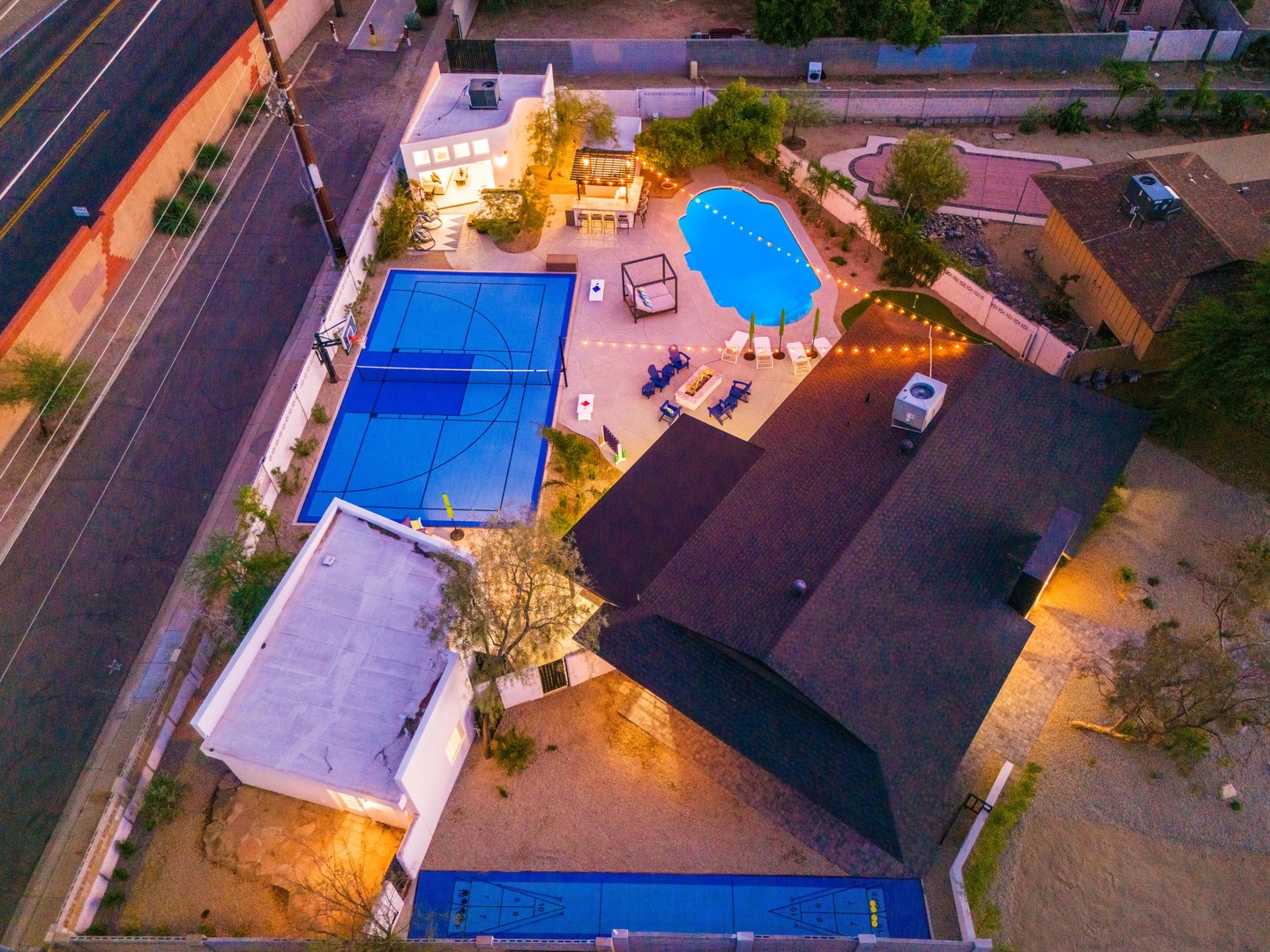 Aerial view of a vacation rental property featuring a large swimming pool, sports court, and outdoor gathering spaces set in a residential neighborhood with convenient street access.