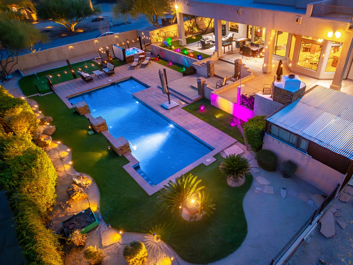 Aerial view of a modern vacation villa featuring a pristine rectangular pool, manicured lawns, and multiple entertainment zones with ambient lighting perfect for evening gatherings. Property includes outdoor dining, lounge areas, and lush tropical landsca