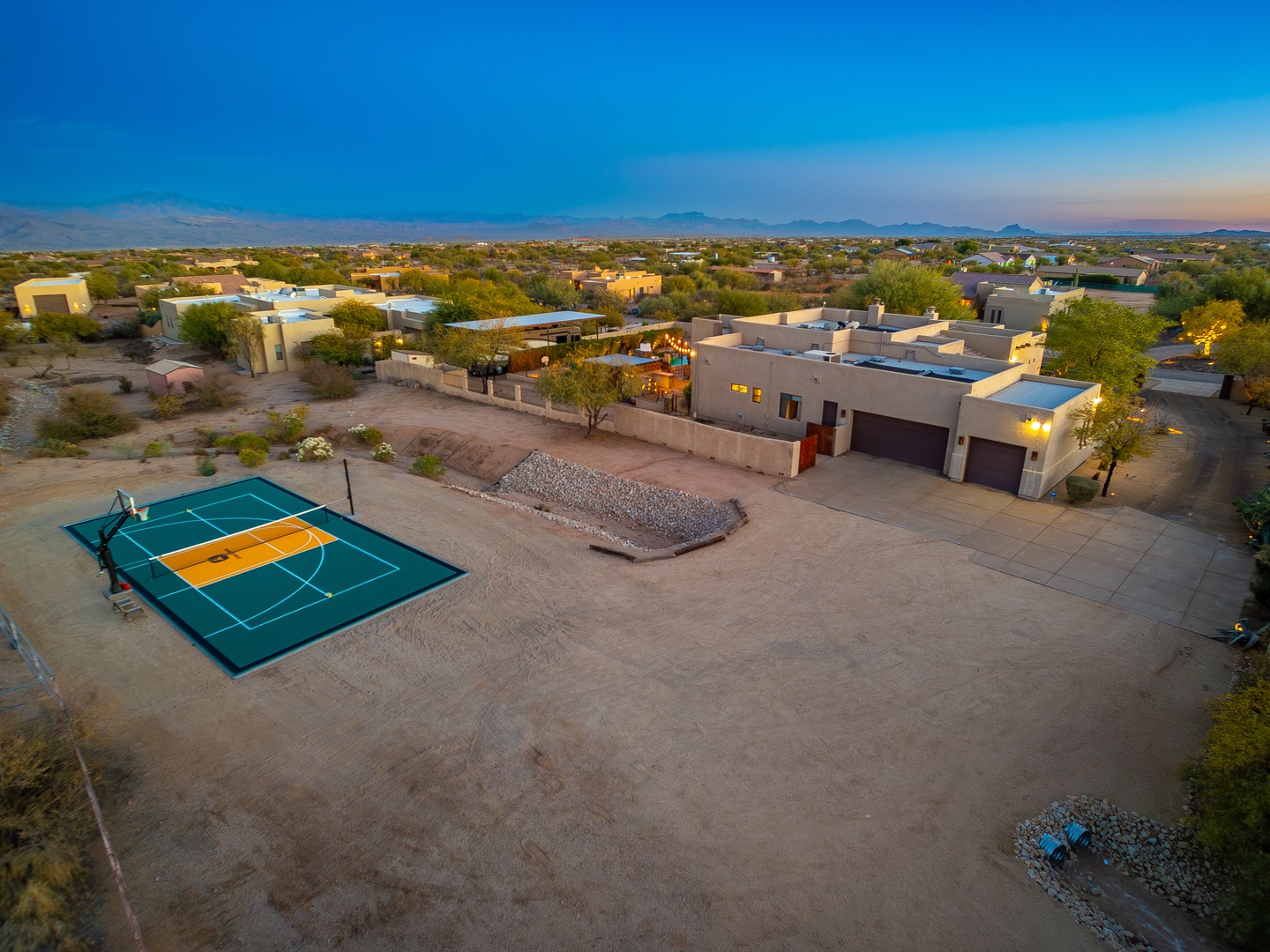 Aerial view of a spacious desert property featuring a full-size sport court, contemporary architecture, and expansive grounds set against mountain vistas and neighborhood surroundings at dusk.