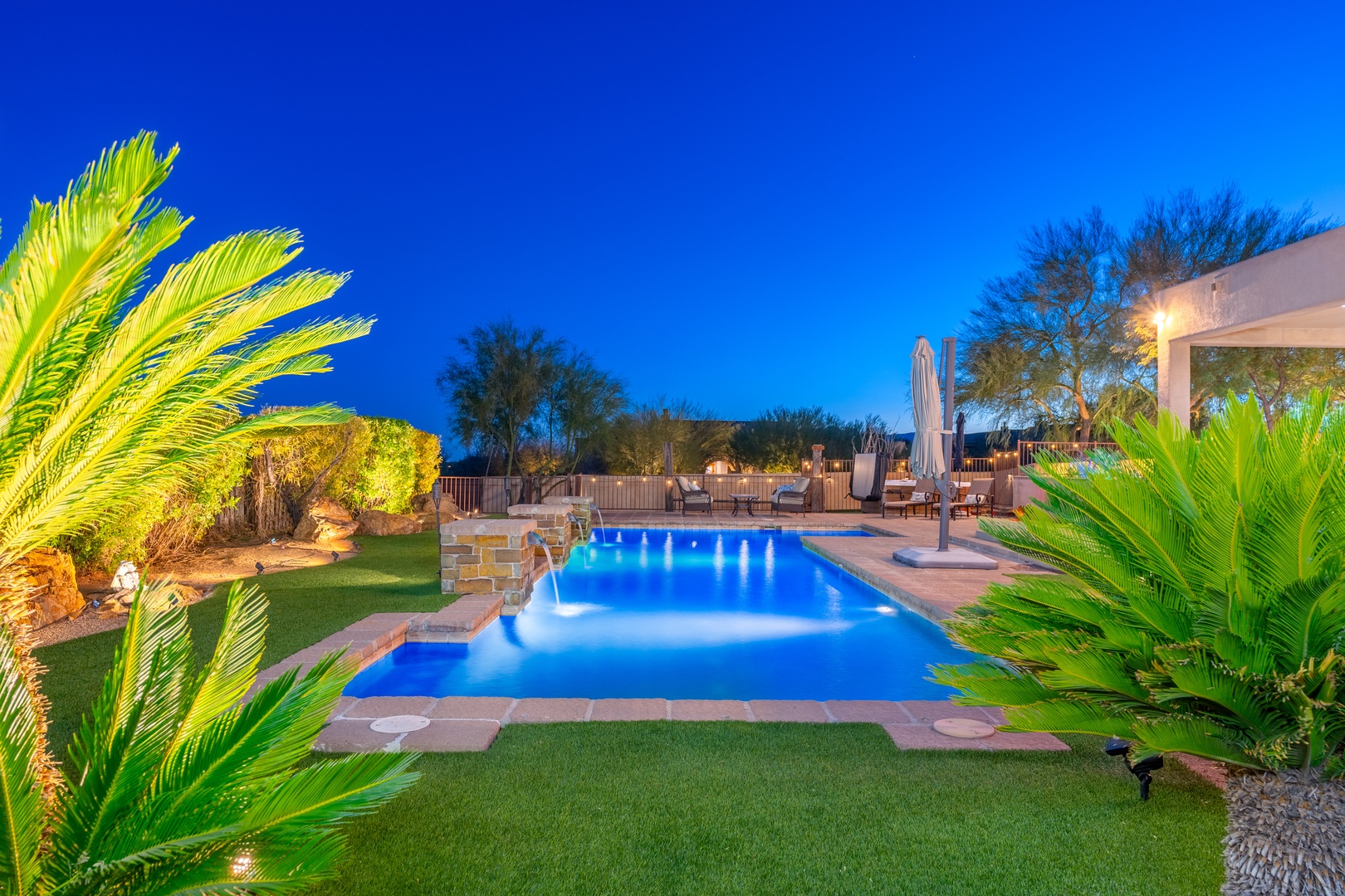 A beautifully lit pool glows under a deep blue evening sky, framed by lush palms and desert landscaping in this resort-style Arizona backyard oasis.