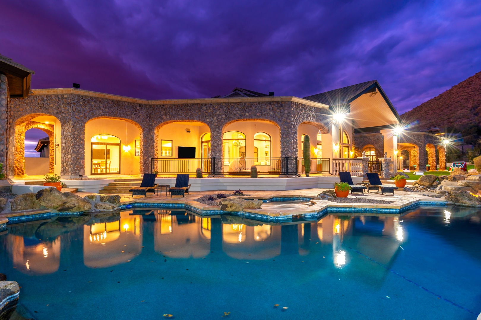Stunning desert estate with illuminated stone archways reflecting in the custom pool beneath dramatic twilight skies and surrounding red rock formations.