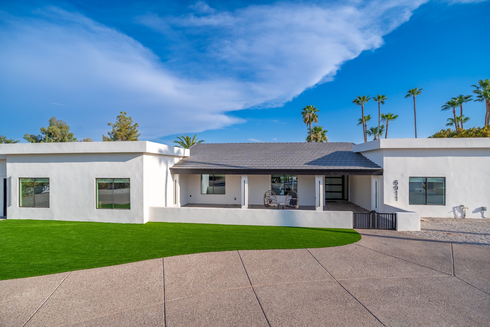 Modern vacation home featuring clean white architecture, lush green lawn, and palm-lined backdrop under bright blue skies—your perfect desert escape awaits.
