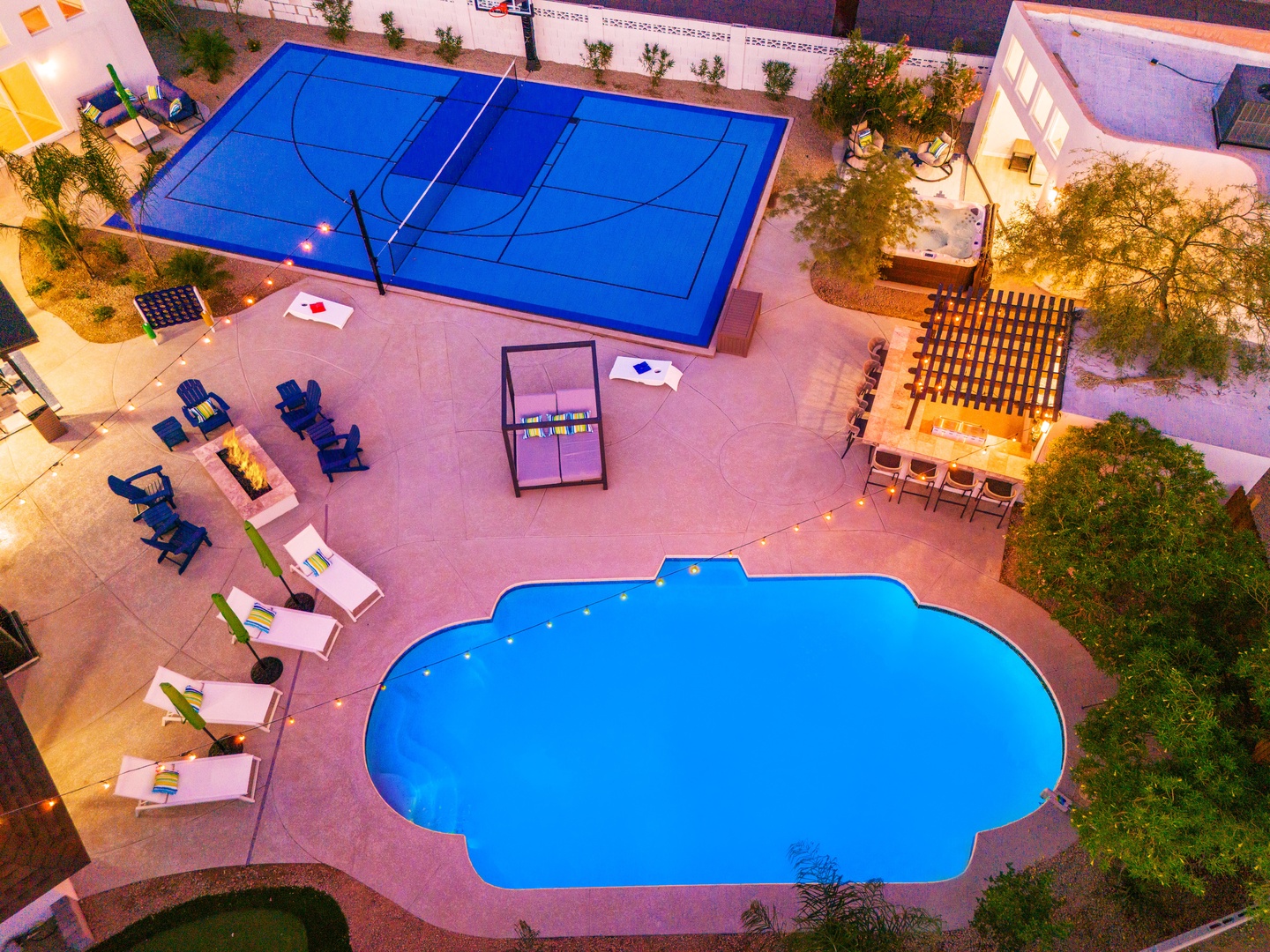 Aerial view showcasing the property's outdoor recreation complex at dusk, featuring a distinctive blue sport court, uniquely shaped swimming pool, multiple seating areas, dining space with pergola, and mature landscaping throughout the courtyard.