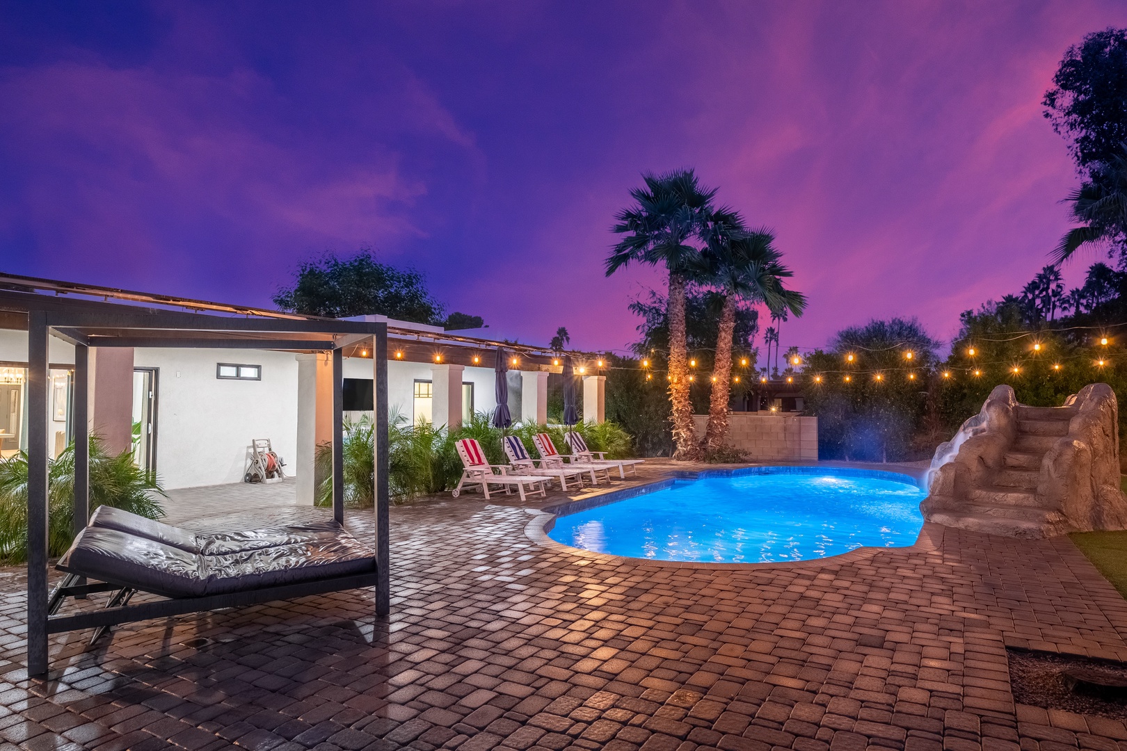 Modern rental home with illuminated pool, lounge chairs, and tropical palms, set beneath a stunning twilight sky perfect for evening relaxation.