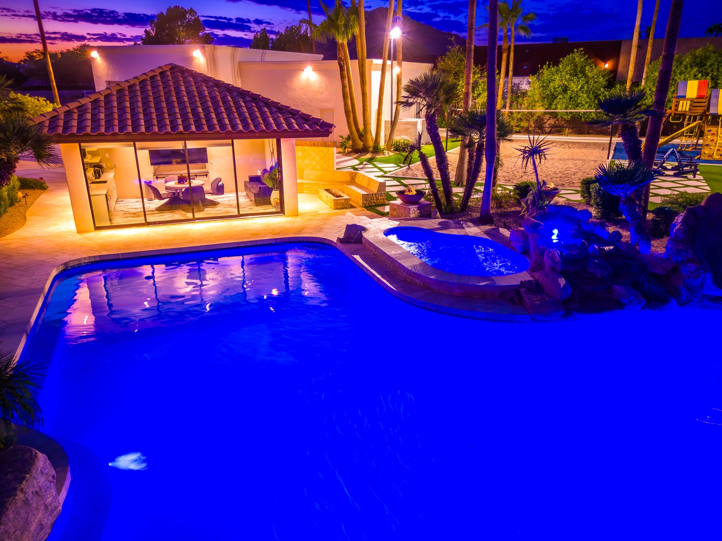 A stunning desert oasis at twilight, featuring a dramatic blue-lit pool, lush landscaping with palms and cacti, and a covered patio area perfect for evening gatherings.
