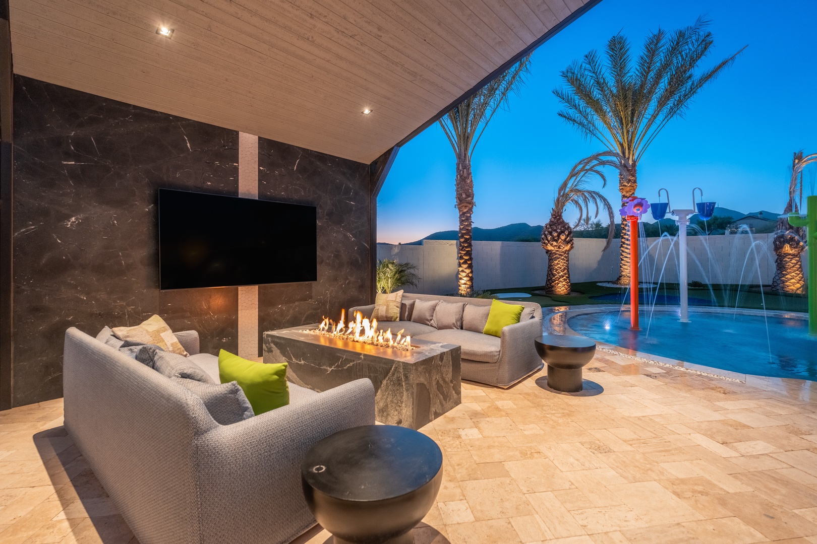 Unwind in your private outdoor oasis with a cozy fireplace, plush seating, and a stunning pool illuminated by colorful fountains under swaying palms and open desert sky.