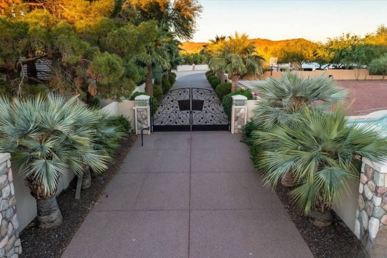 A gated entrance leads to this desert retreat, framed by lush palms and set against golden hillsides bathed in warm evening light—a welcoming arrival experience.