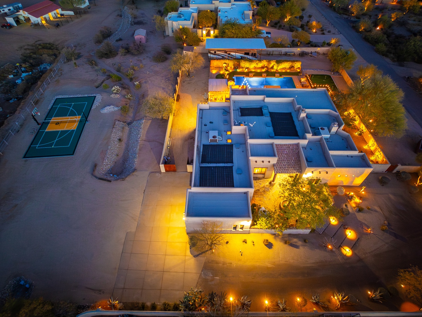 Aerial view of a stunning southwestern-style property with illuminated courtyard spaces, multiple pools, tennis court, and desert landscaping—a luxurious desert retreat offering exceptional private amenities.