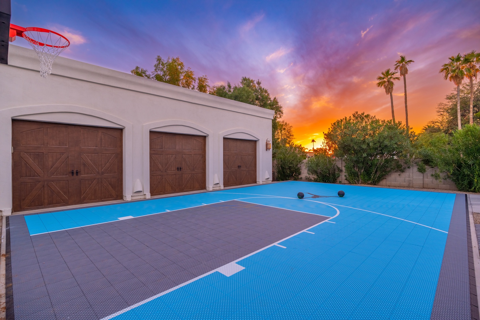 The property features a private outdoor basketball court framed by striking sunsets, desert palms, and lush greenery—perfect for recreation near ample garage space.