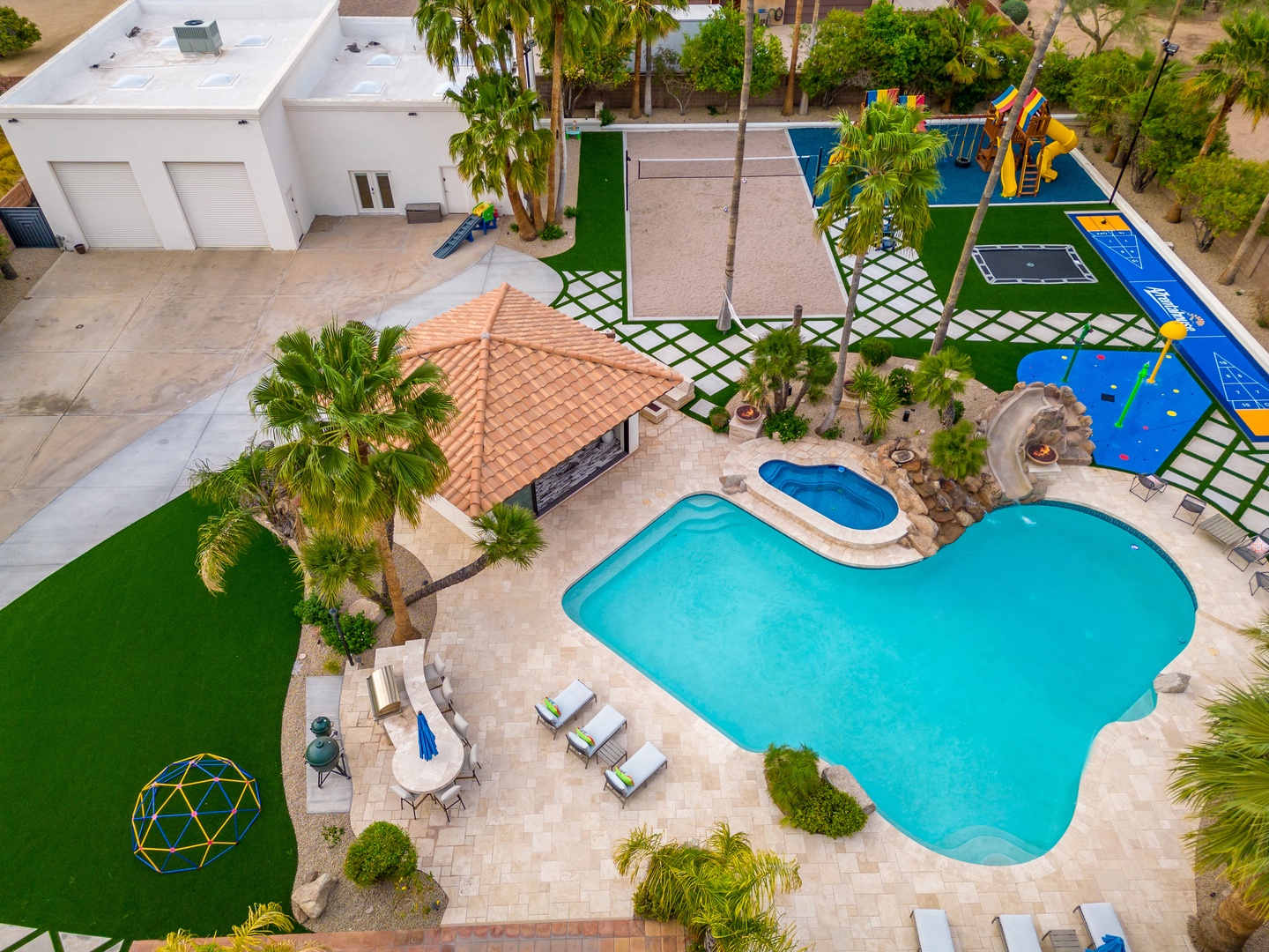 Aerial view reveals a private resort-style backyard with a sprawling freeform pool, intimate spa, covered pavilion, lush palm landscaping, and dedicated children's play areas—an expansive outdoor retreat for groups and families.