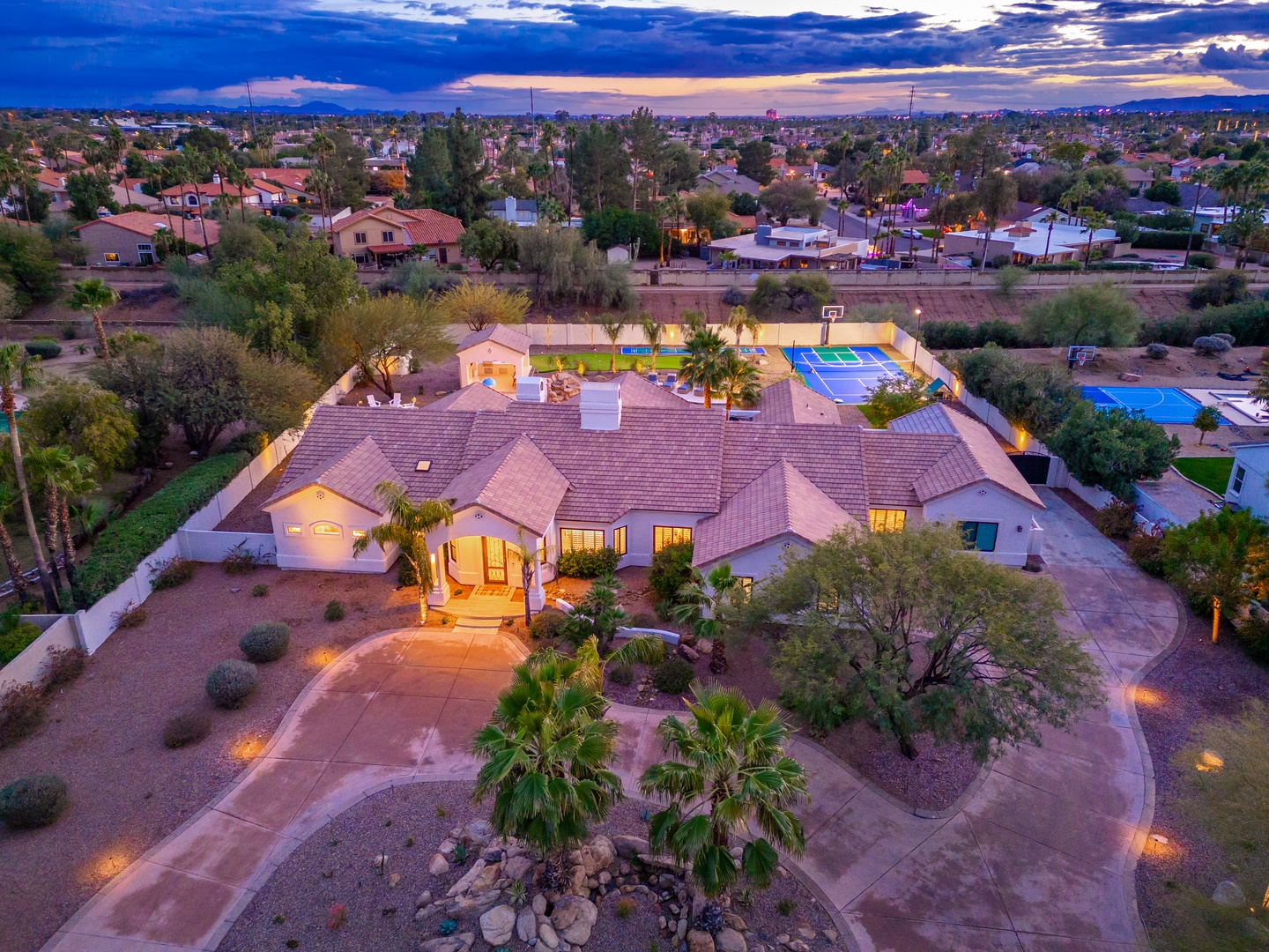 Aerial view of a spacious residential property in a suburban neighborhood, featuring a Mediterranean-style home with a tiled roof, private pool, tennis court, and beautifully landscaped grounds illuminated by pathway lighting at dusk.