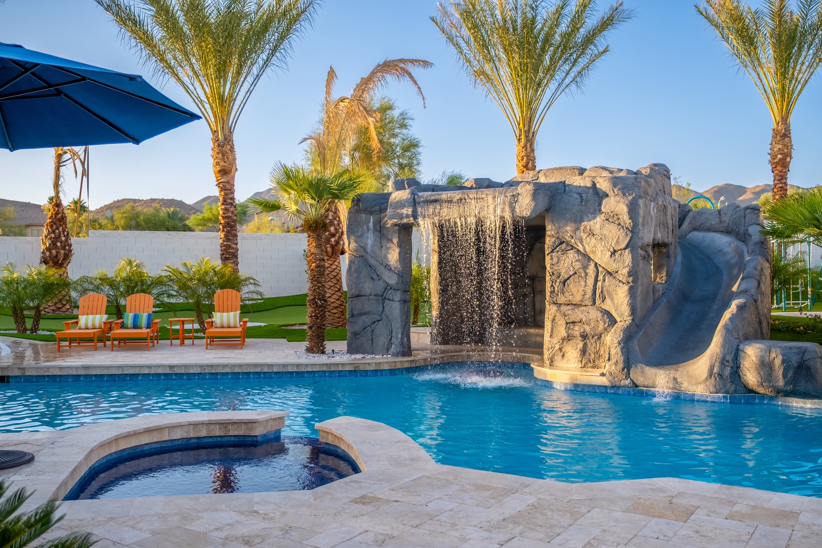 A stunning resort-style pool features a dramatic stone waterfall, sparkling hot tub, and lounge chairs beneath swaying palms—your private oasis awaits under desert skies.