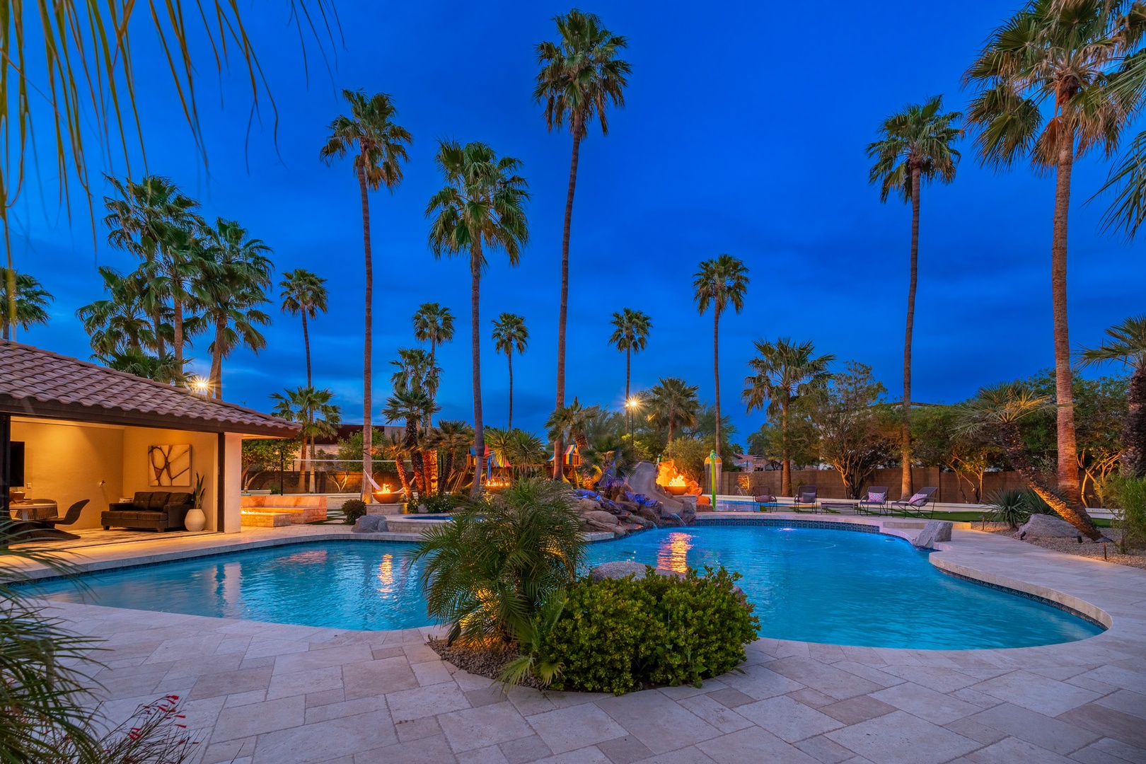 A stunning resort-style pool area nestled beneath swaying palms and a deep blue evening sky, featuring illuminated water features, natural stone accents, and lush tropical landscaping perfect for relaxing under the stars.