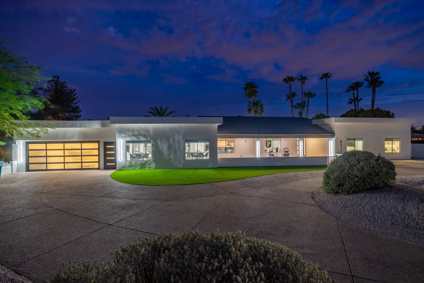 A striking modern desert retreat illuminated at twilight, surrounded by manicured lawns and towering palms in a tranquil neighborhood.