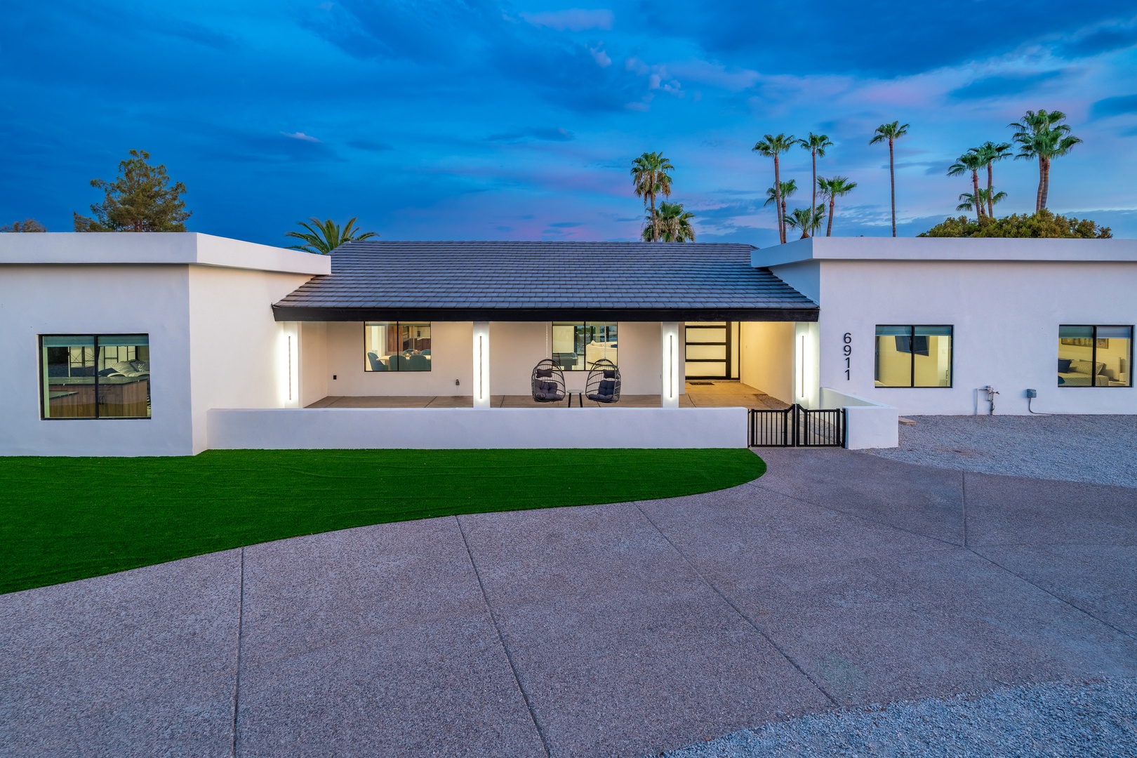Modern single-story property with palm tree backdrop, featuring expansive driveway and beautifully landscaped lawn beneath desert twilight sky.