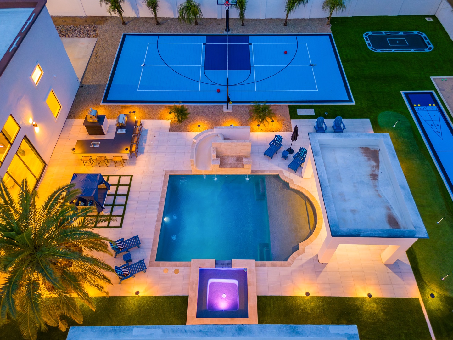 Aerial view of a modern vacation rental featuring a central pool, basketball court, outdoor dining patio, and multiple recreation areas, all illuminated for evening enjoyment.