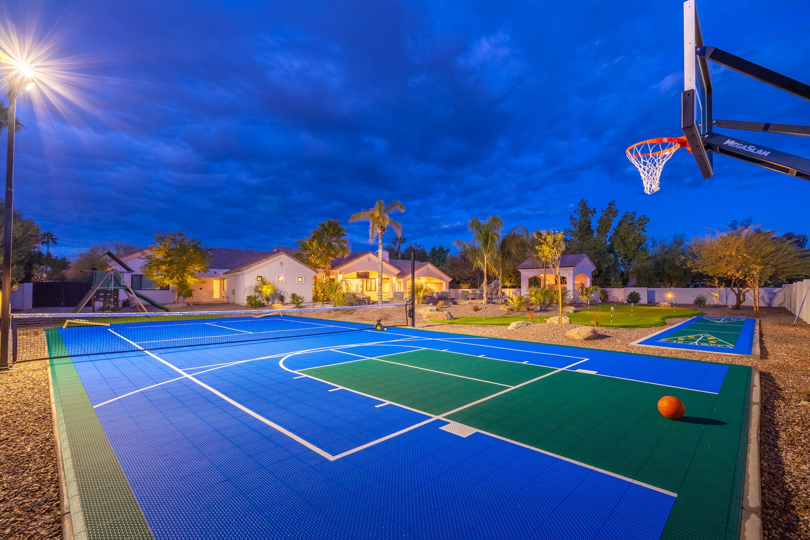 The property features a private multi-sport court illuminated for evening play, surrounded by lush landscaping and mature palm trees in a peaceful residential setting.