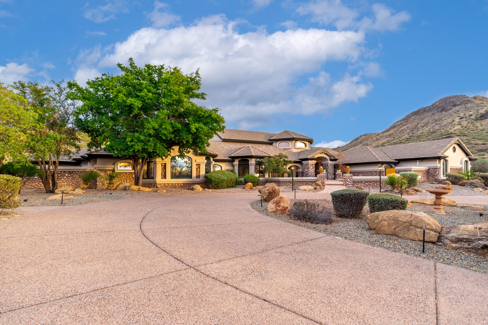 Expansive desert estate nestled against mountain foothills, featuring Mediterranean-style architecture, mature shade trees, and meticulously landscaped grounds with natural stone accents throughout the property.