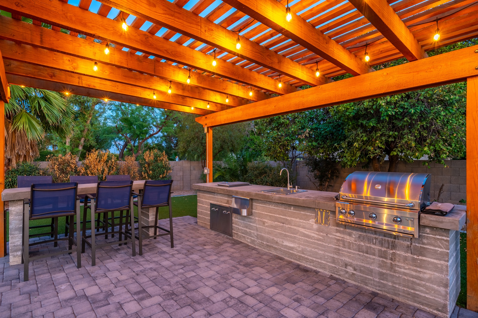 Gather around your private outdoor kitchen where warm string lights and a built-in grill create the perfect setting for evening meals under a beautiful pergola.