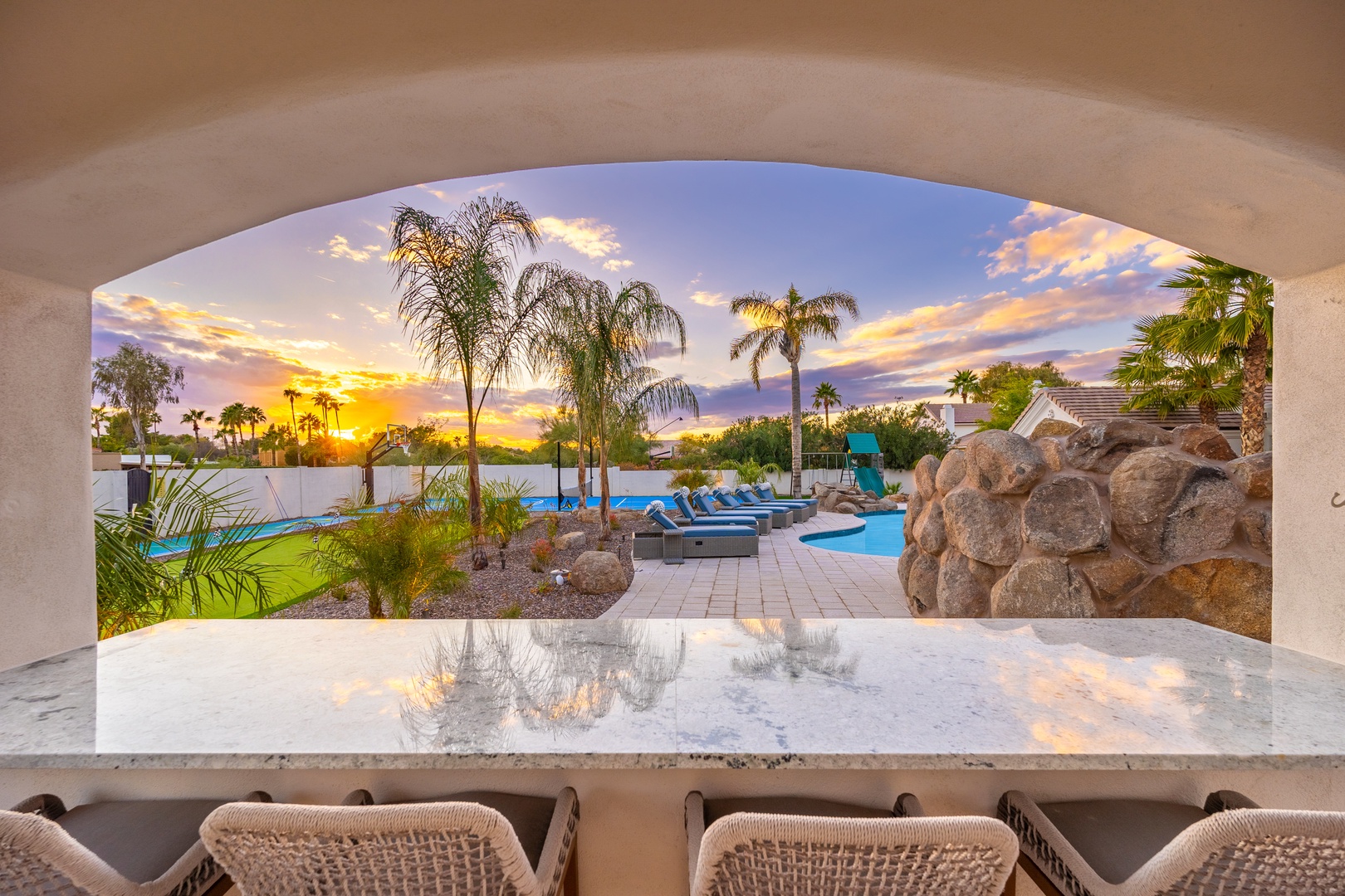 Golden hour paints the sky beyond the covered patio, framing lush palms, a sparkling pool, and loungers awaiting sunset cocktails.