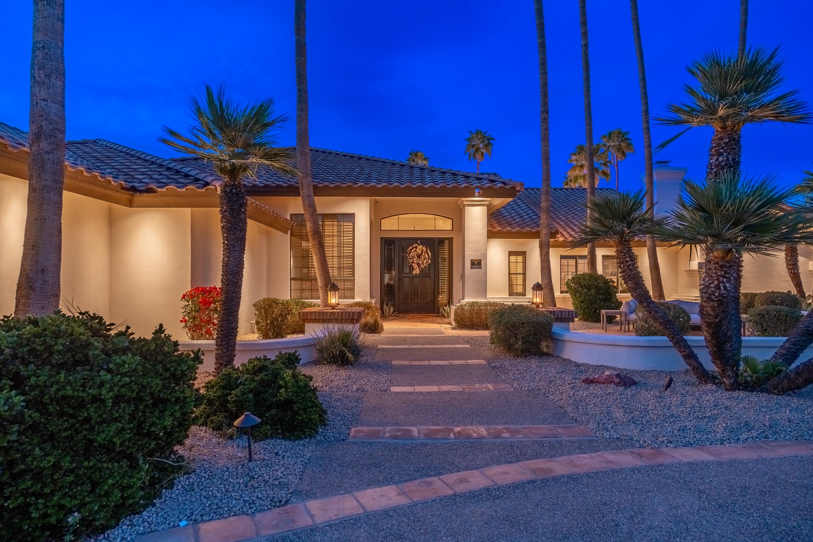 A stunning desert oasis property with elegant Mediterranean architecture, framed by towering palm trees and desert landscaping, perfectly lit as evening falls.