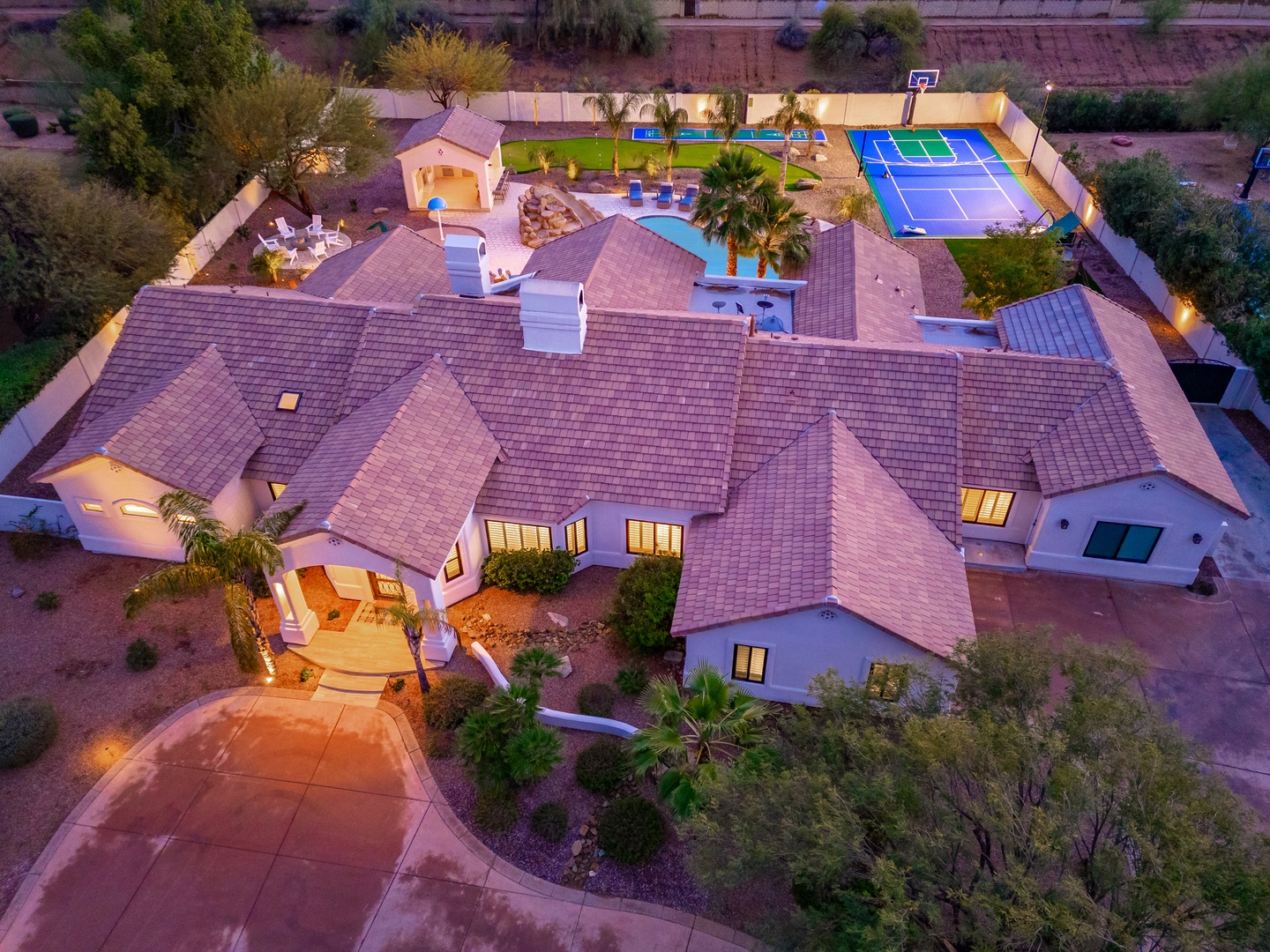 Aerial view of an expansive vacation rental estate featuring a private swimming pool, full tennis court, basketball hoop, covered outdoor entertaining spaces, and manicured desert landscaping with palm trees and lush lawns.