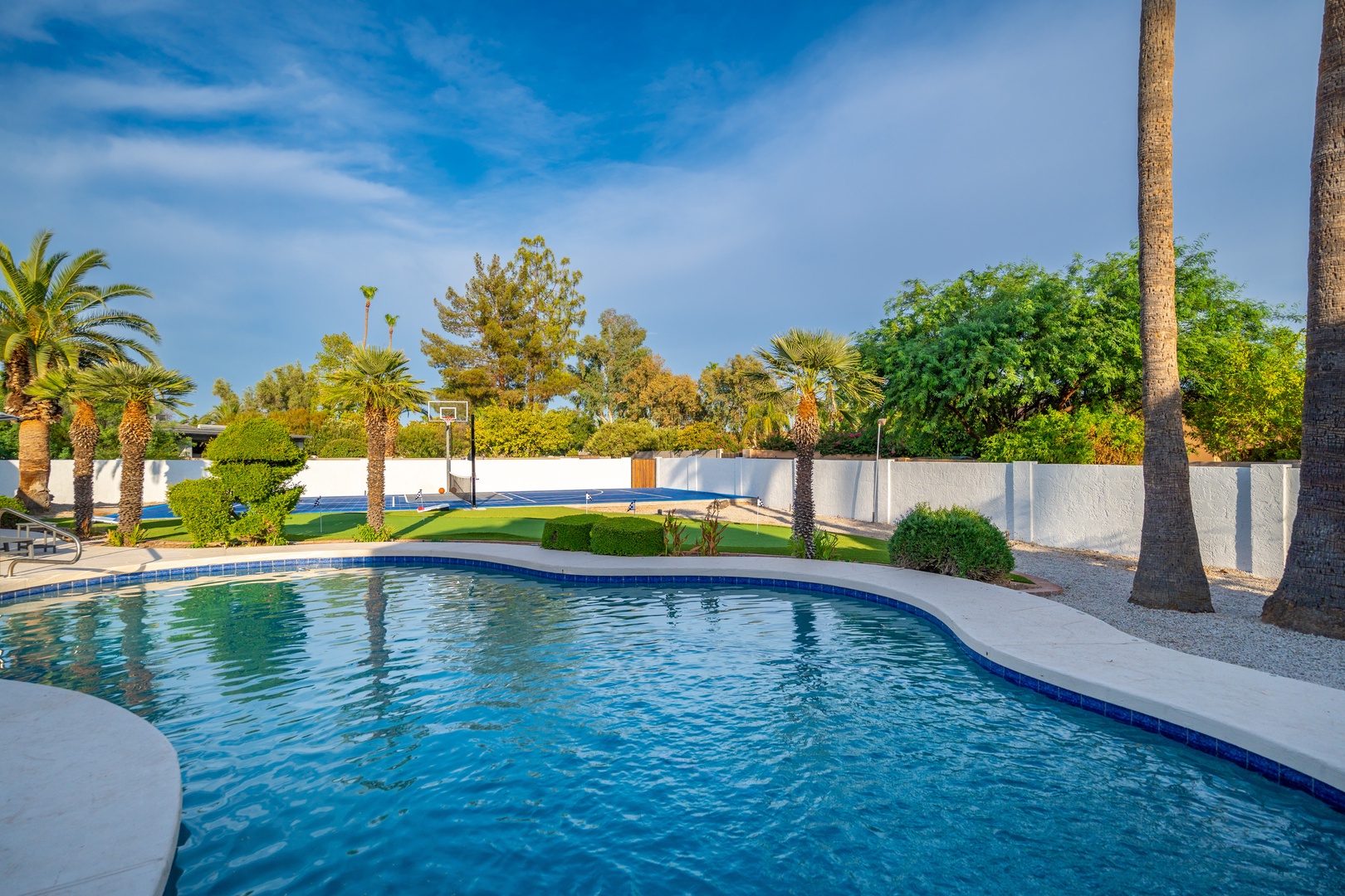 A sparkling pool awaits, framed by palms and a private basketball court. Dive into the blue waters or lounge on deck surrounded by lush greenery under sunny skies.