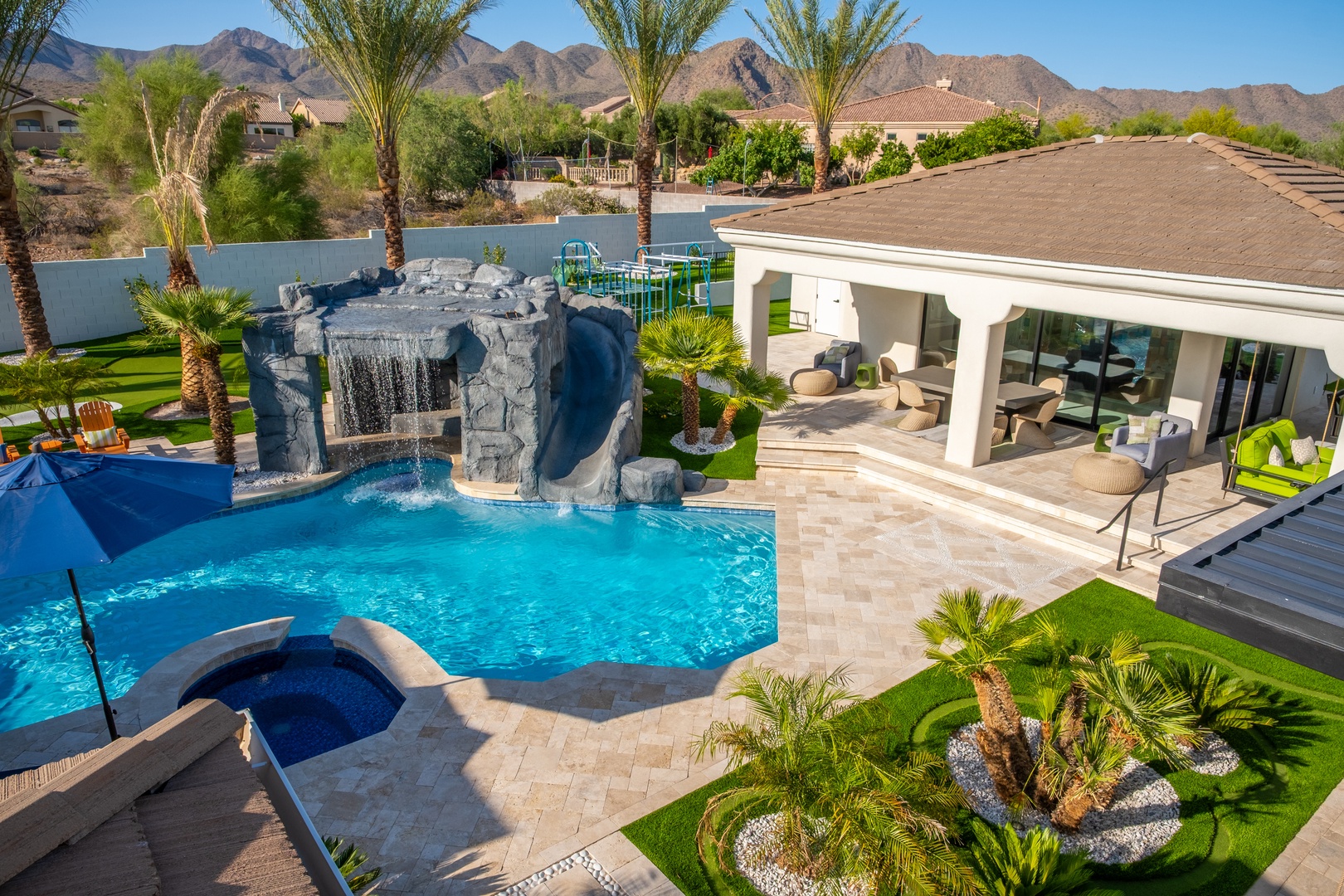 The property features a resort-style pool with a dramatic waterfall grotto and water slide, surrounded by lush landscaping with palm trees, covered patio seating areas, and mountain views.
