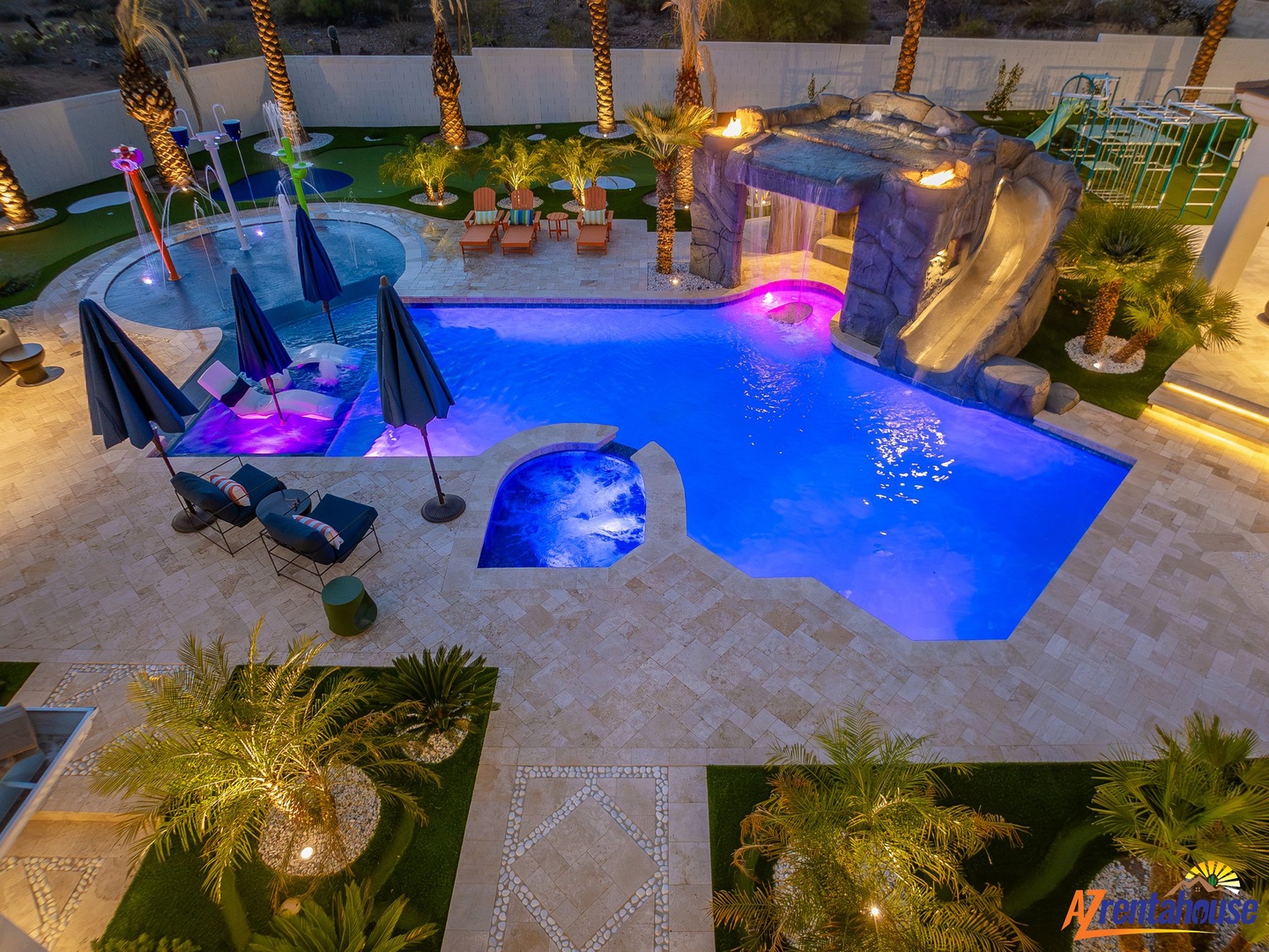 Spectacular resort-style pool complex with water slide, hot tub, and tropical landscaping illuminated for evening enjoyment.