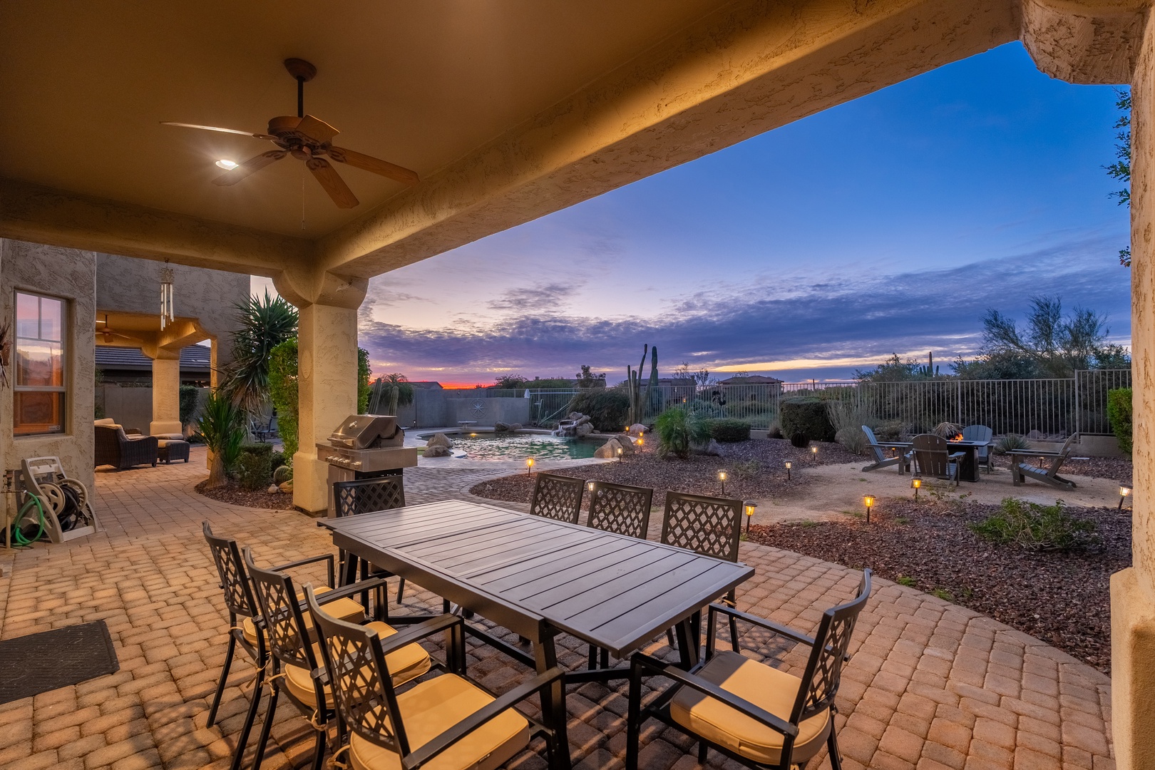 A covered patio with desert landscaping captures the vibrant evening sky. The property features BBQ facilities, outdoor dining, and a serene desert setting perfect for relaxation.