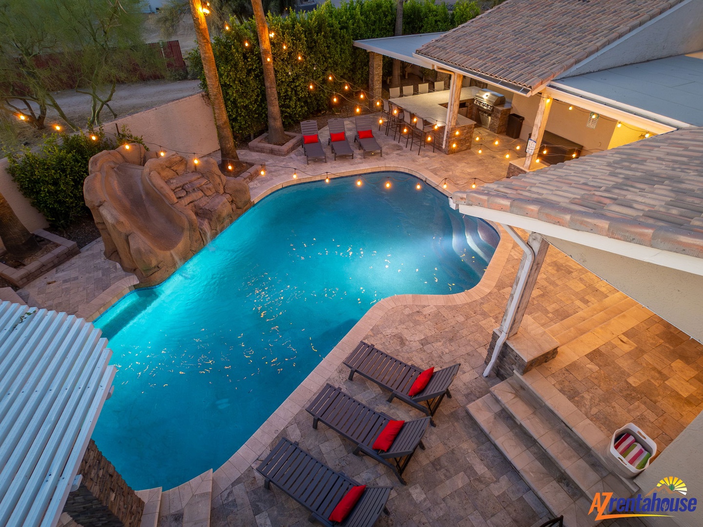 Elevated aerial view showcasing the property's inviting pool area with integrated rock waterfall, covered outdoor kitchen and bar, ambient string lighting, and multiple seating zones creating an ideal entertainment space.