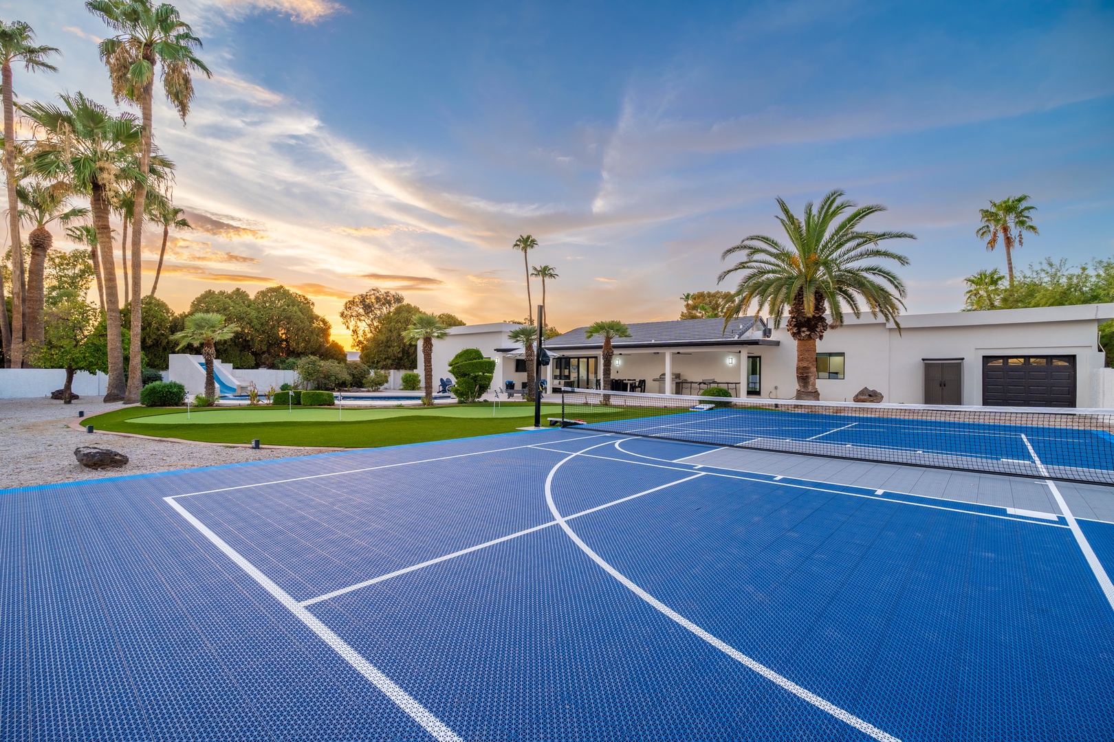 A modern property with sport court, lush lawn, and palm trees set against a stunning sunset sky. The spacious grounds feature multiple recreational areas perfect for outdoor activities.