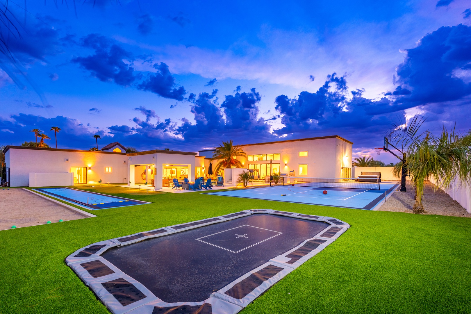 A stunning desert oasis at twilight featuring a spacious pool, sports court, and vibrant landscaping under dramatic evening skies. The warm glow of the home invites relaxation and outdoor fun.