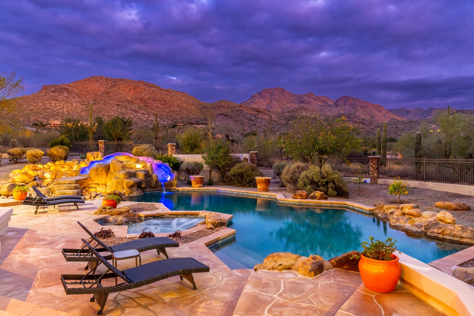 Dramatic desert mountains provide a stunning backdrop to this resort-style pool with waterslide and integrated hot tub, captured during a beautiful twilight hour.