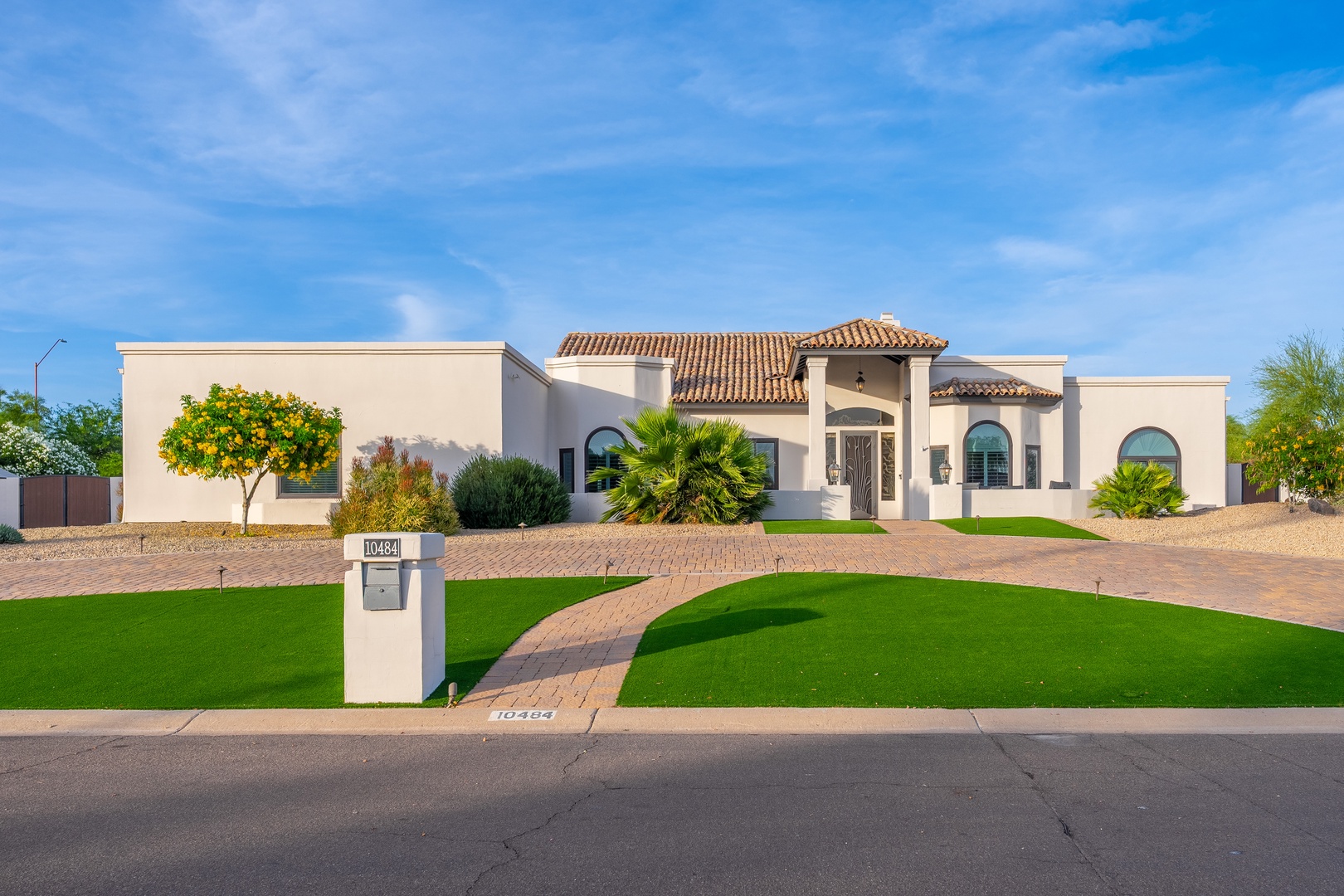 Mediterranean-style vacation rental with terracotta tile roof, arched windows, and manicured desert landscaping featuring palm trees and lush green lawn. Located in a quiet residential neighborhood with ample driveway parking.