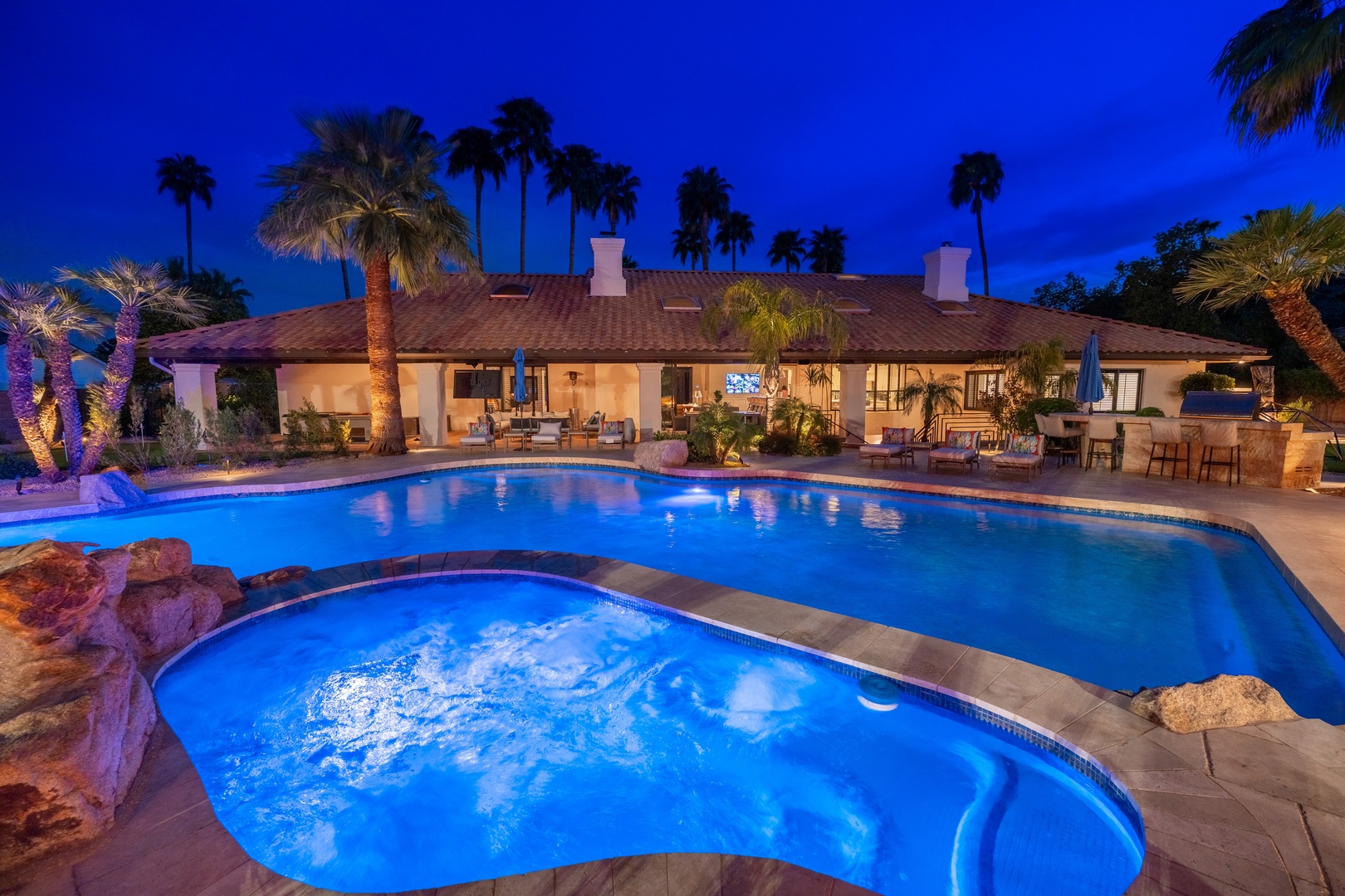 Luxurious private oasis featuring a sparkling pool and hot tub surrounded by towering palms, with elegant outdoor dining and lounge areas perfect for evening relaxation under the desert sky.