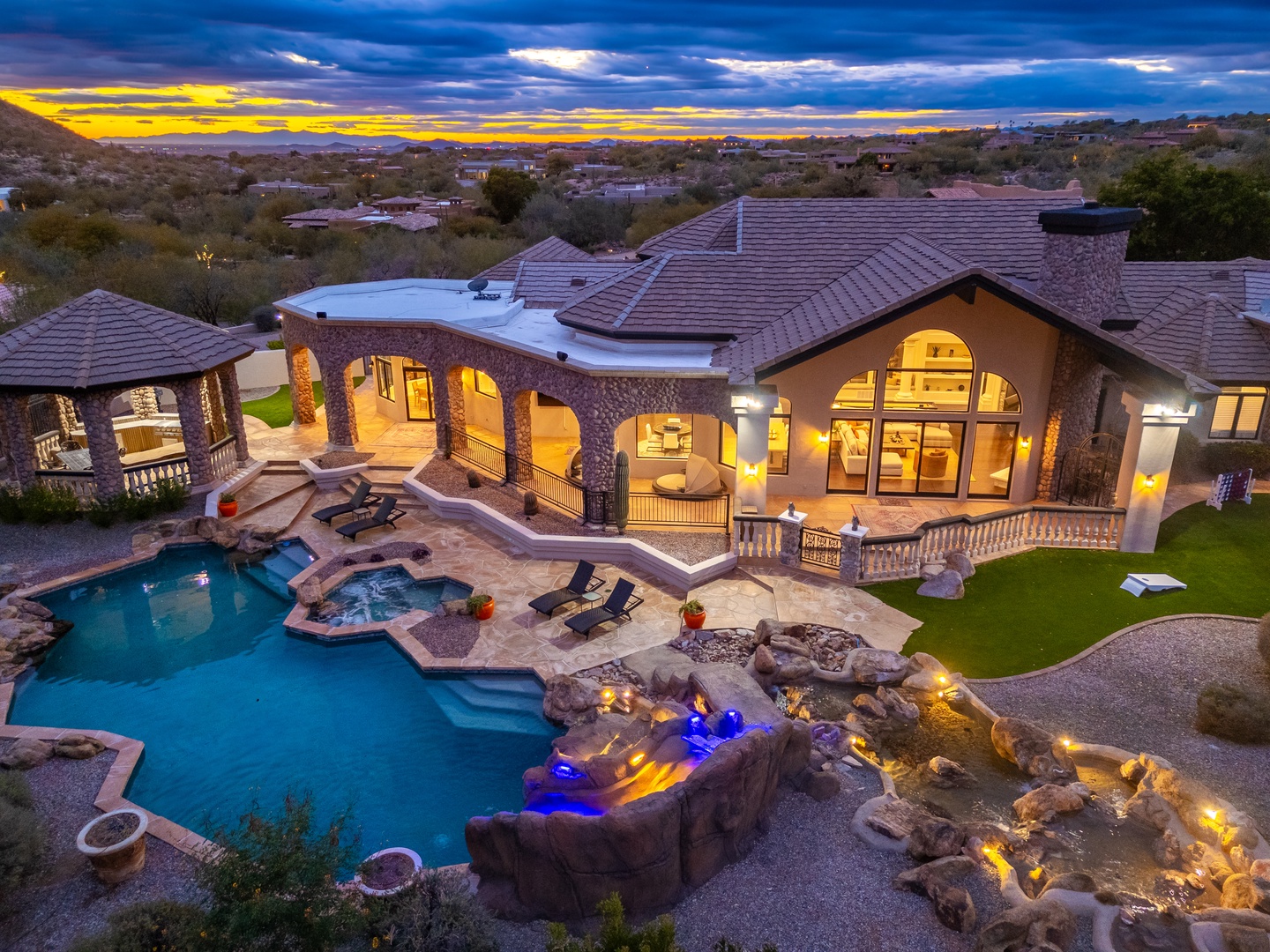 Spectacular desert estate at twilight, featuring an expansive stone villa with a custom pool, grotto-style hot tub, and waterfall features set against a dramatic Arizona sunset and distant mountain views.