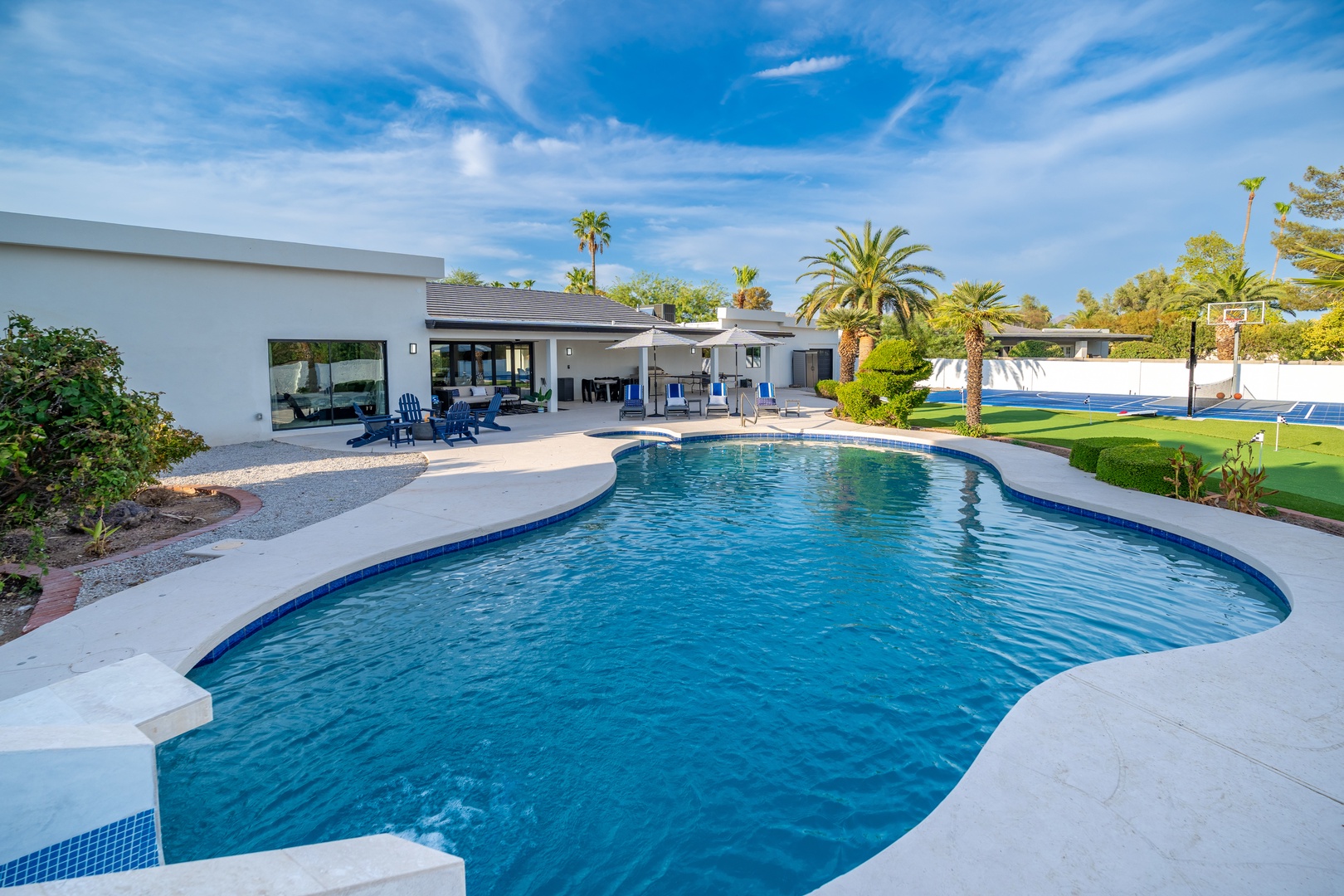 A stunning resort-style pool stretches across the backyard, surrounded by expansive lawn and palm trees, with covered patio seating, sports courts, and mountain views completing this private oasis.
